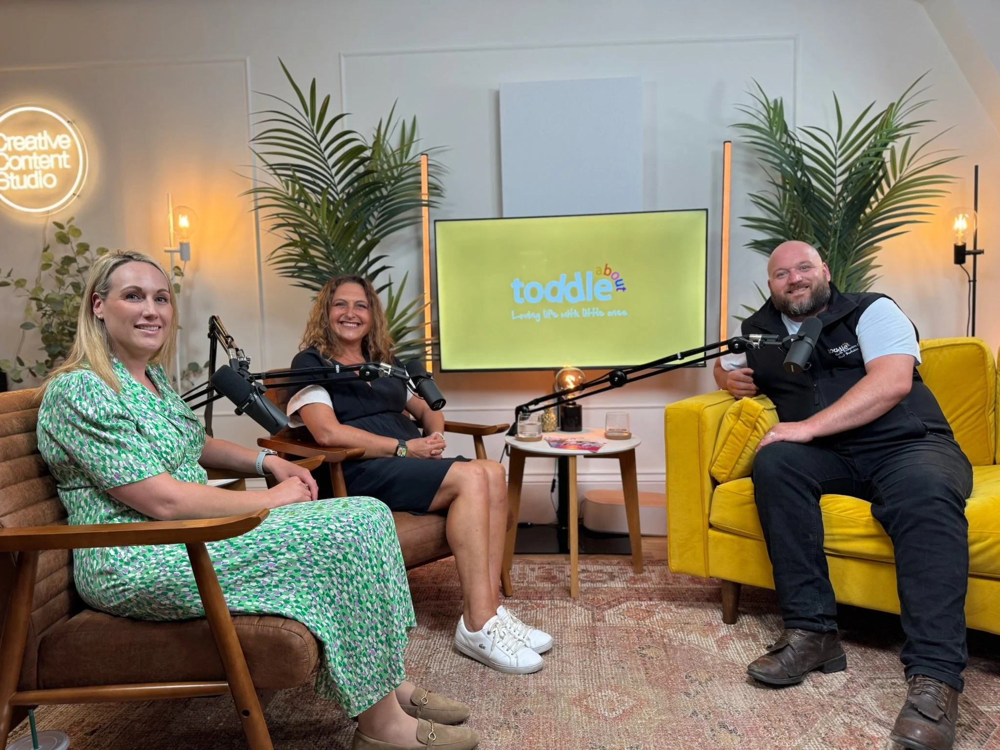 Three people seated on couches in a studio with a television screen displaying the logo 'Toddle about.' The woman on the left is wearing a green patterned dress, the woman in the middle is wearing a black dress with white sneakers, and the man on the
