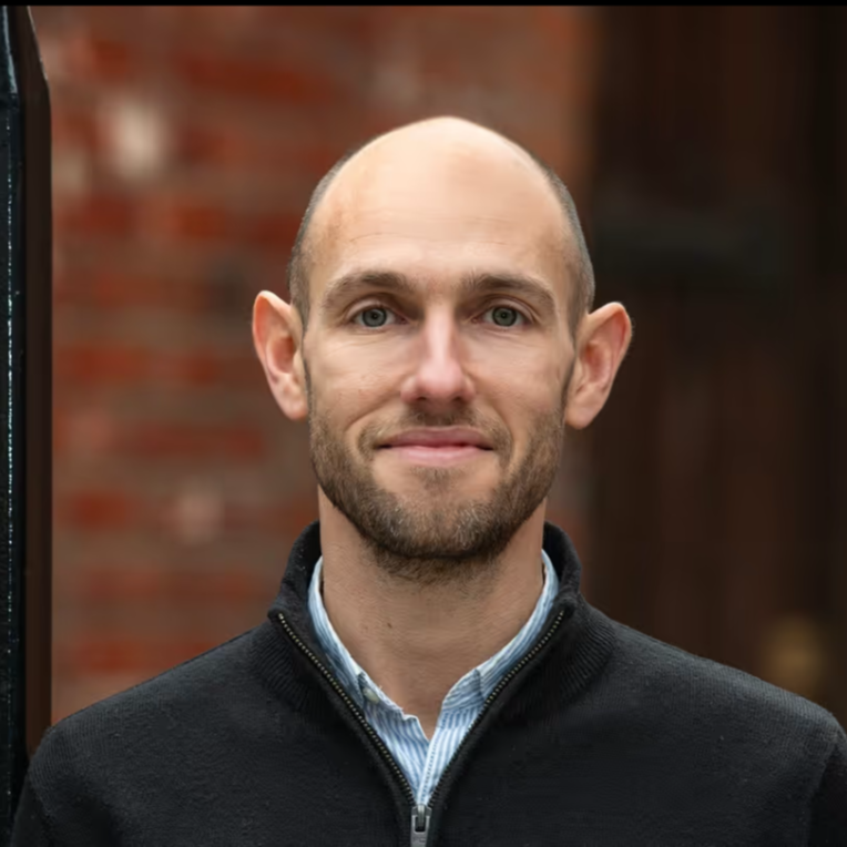 Rich Webly, marketing consultant  wearing a black zip-up jacket and a light blue collared shirt, standing outdoors with a blurred brick wall background.
