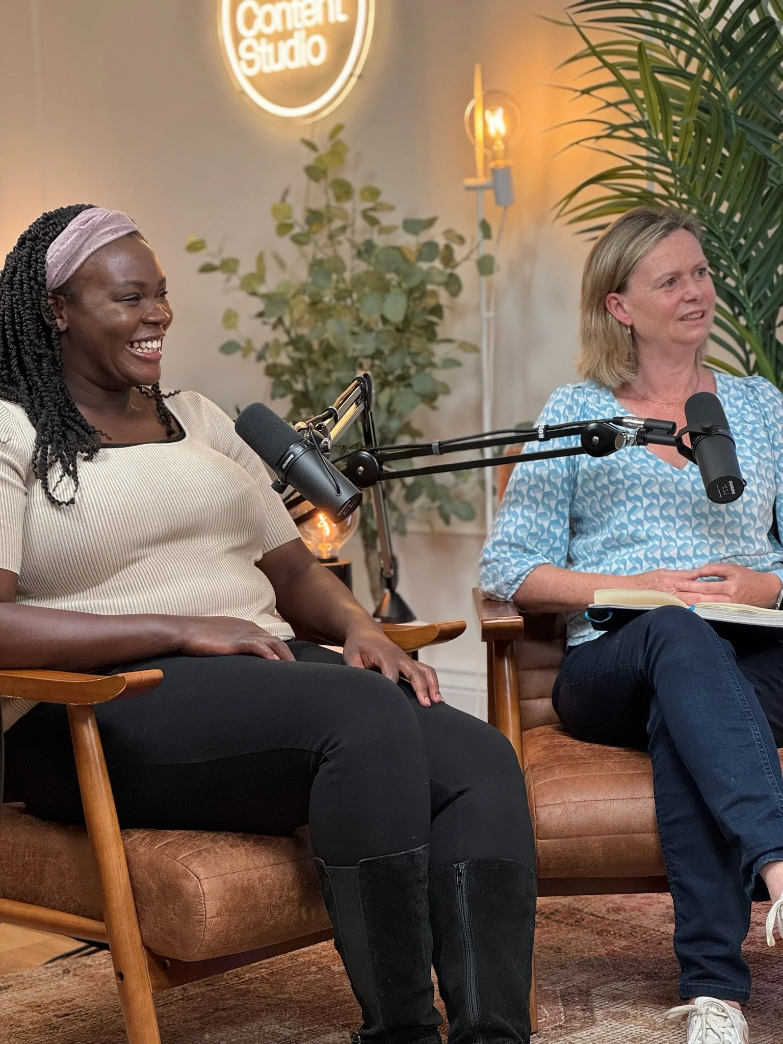 Two women sitting on chairs in a recording studio, smiling and speaking into microphones. Background includes a plant, a neon sign reading 'Content Studio,' and warm ambient lighting.