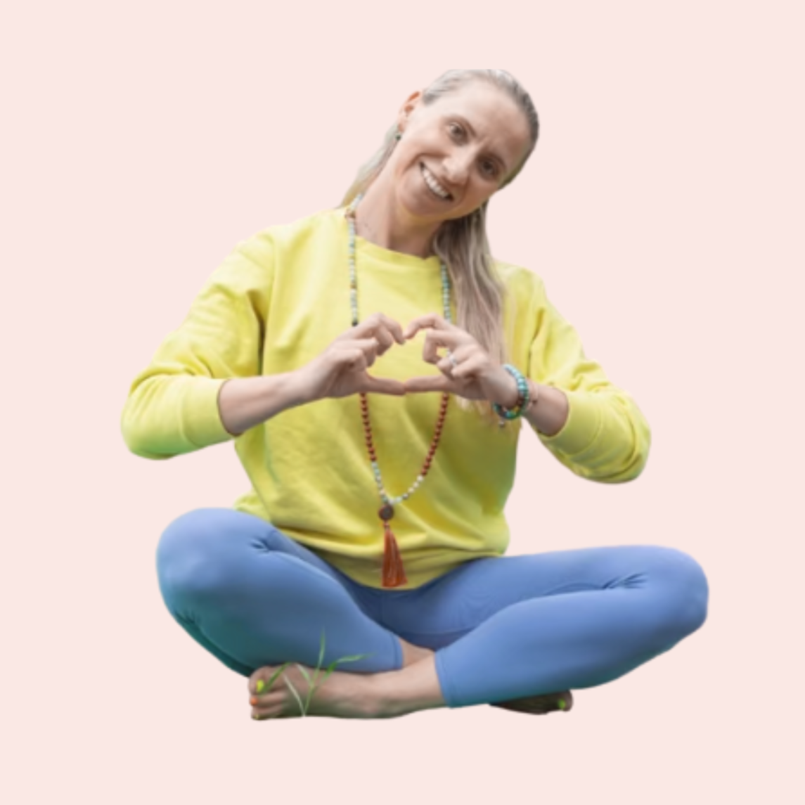 A woman sitting cross-legged on the floor, wearing a yellow sweatshirt and blue leggings, making a heart shape with her hands, smiling, against a light pink background.