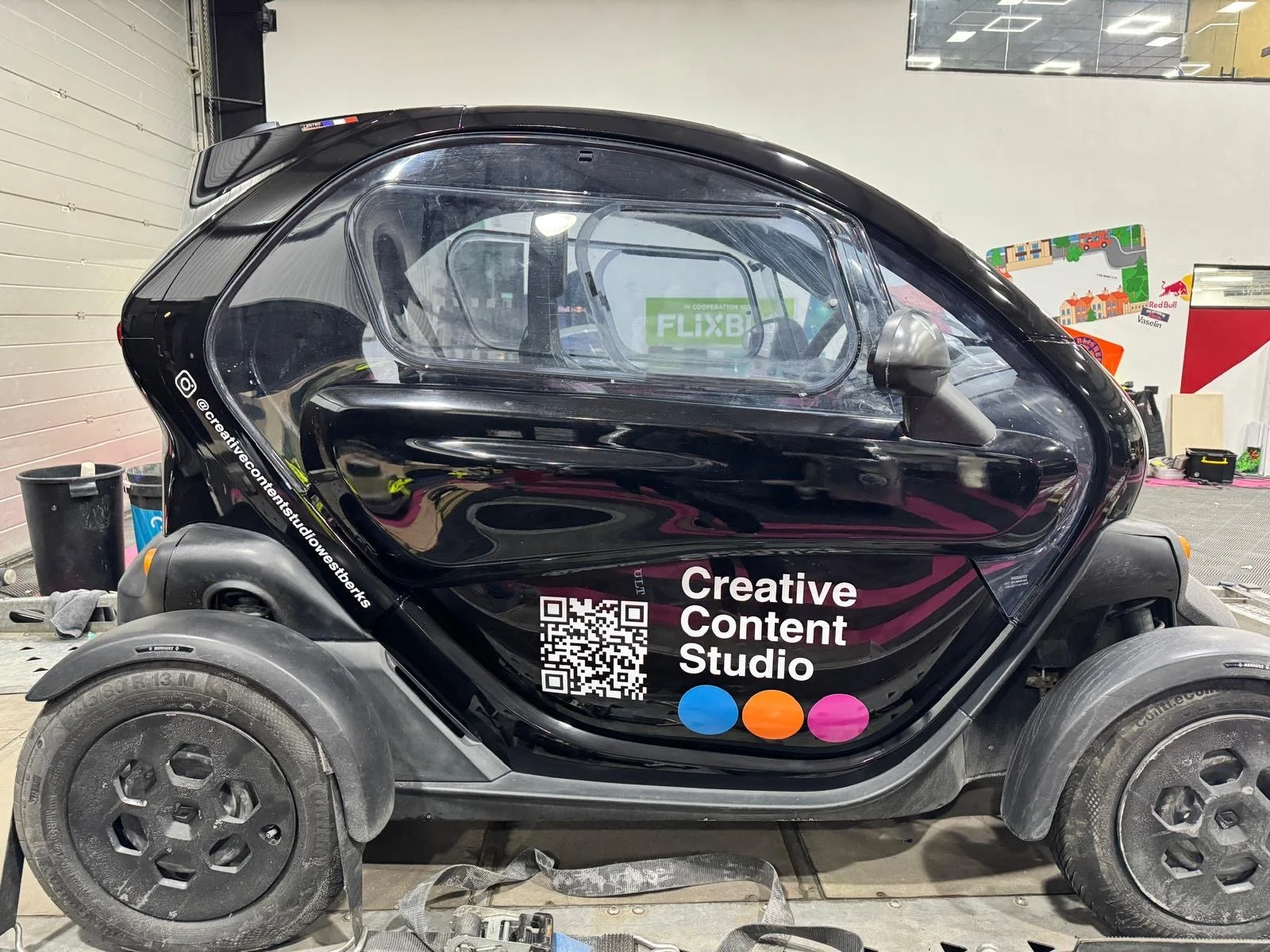 A black enclosed small electric vehicle with transparent canopy and sticker that reads "Creative Content Studio" and a QR code on the side, located indoors in a workshop or garage.