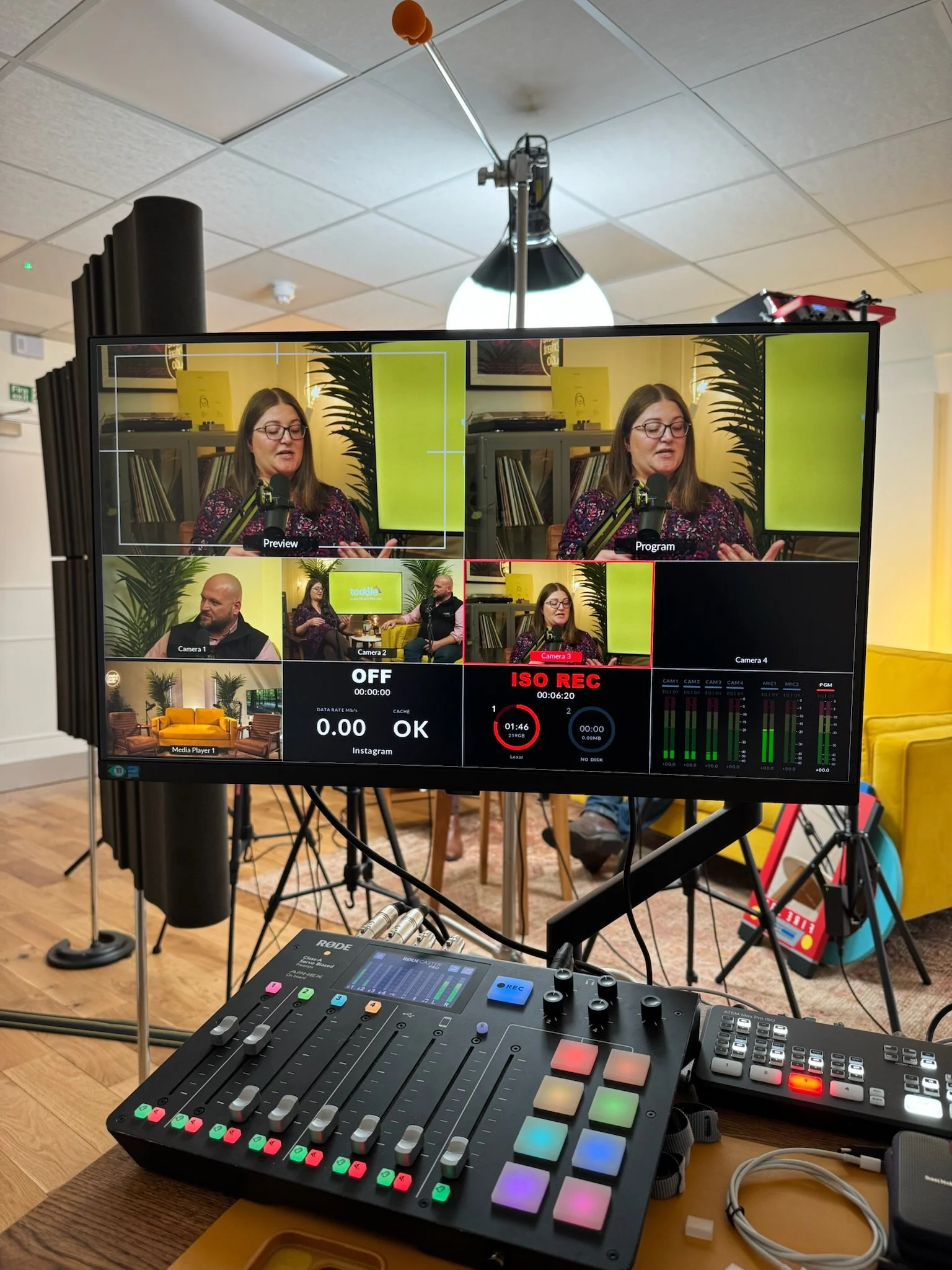 A video production monitor displays a multi-camera interview setup where a woman is speaking, with other camera angles shown. A control panel and audio equipment are in the foreground, and a woman is seen speaking via microphone in a studio setting w