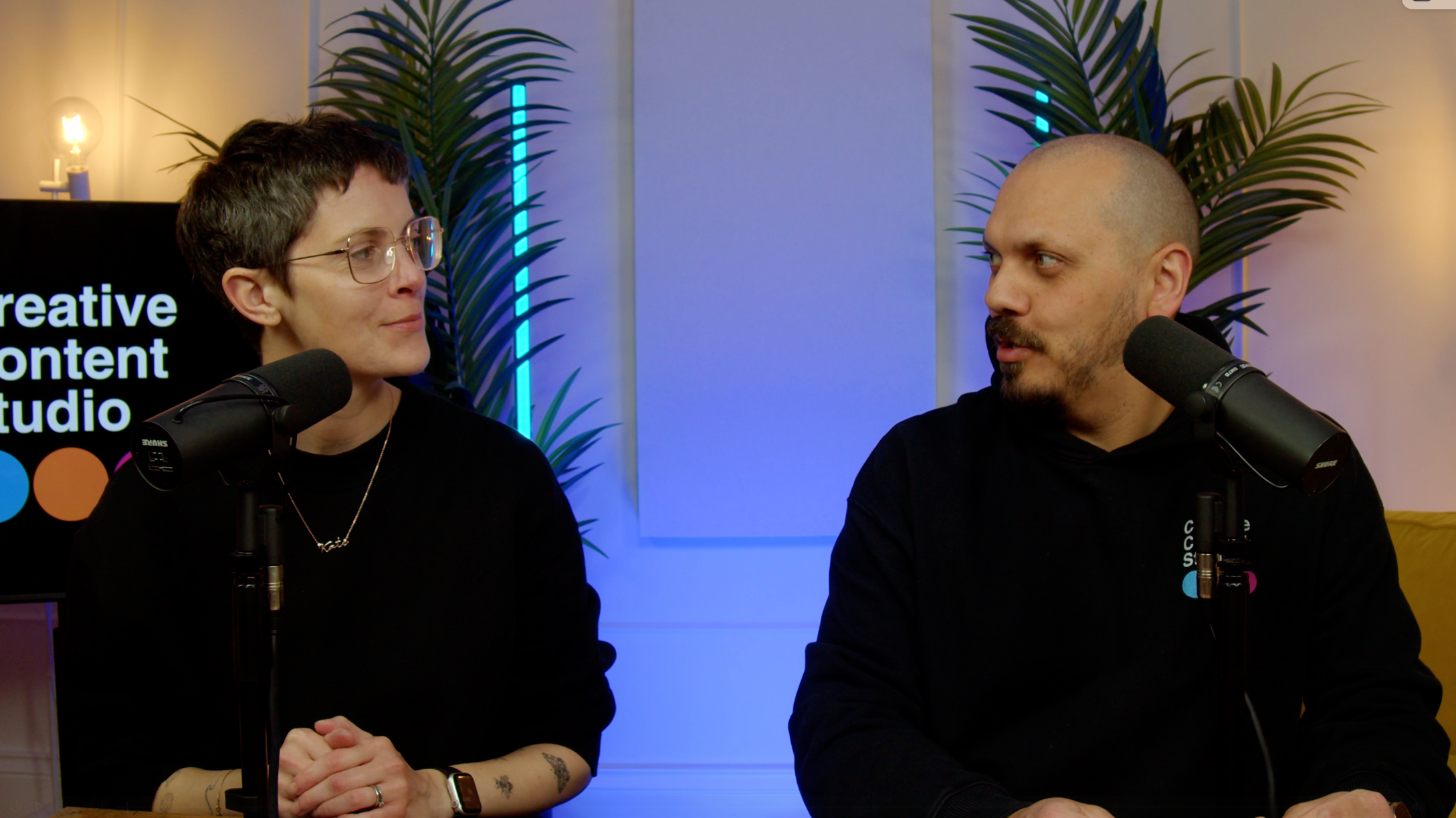 Two people sitting at a table with microphones, engaging in conversation, surrounded by indoor plants and colorful ambient lighting.