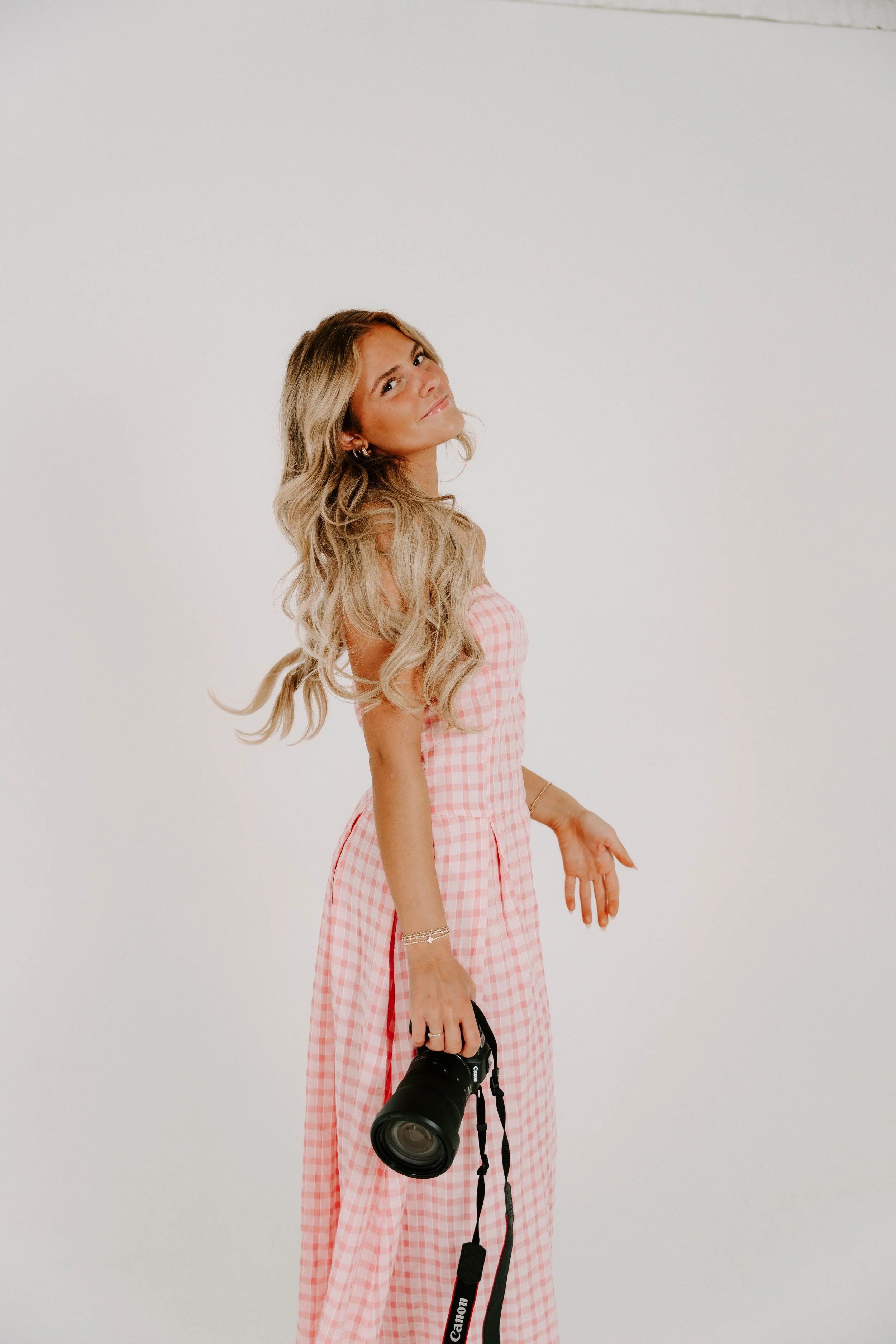 A young woman with blonde hair wearing a polka dot dress, white sneakers, and a black jacket draped over her shoulders standing in a minimalist white room.