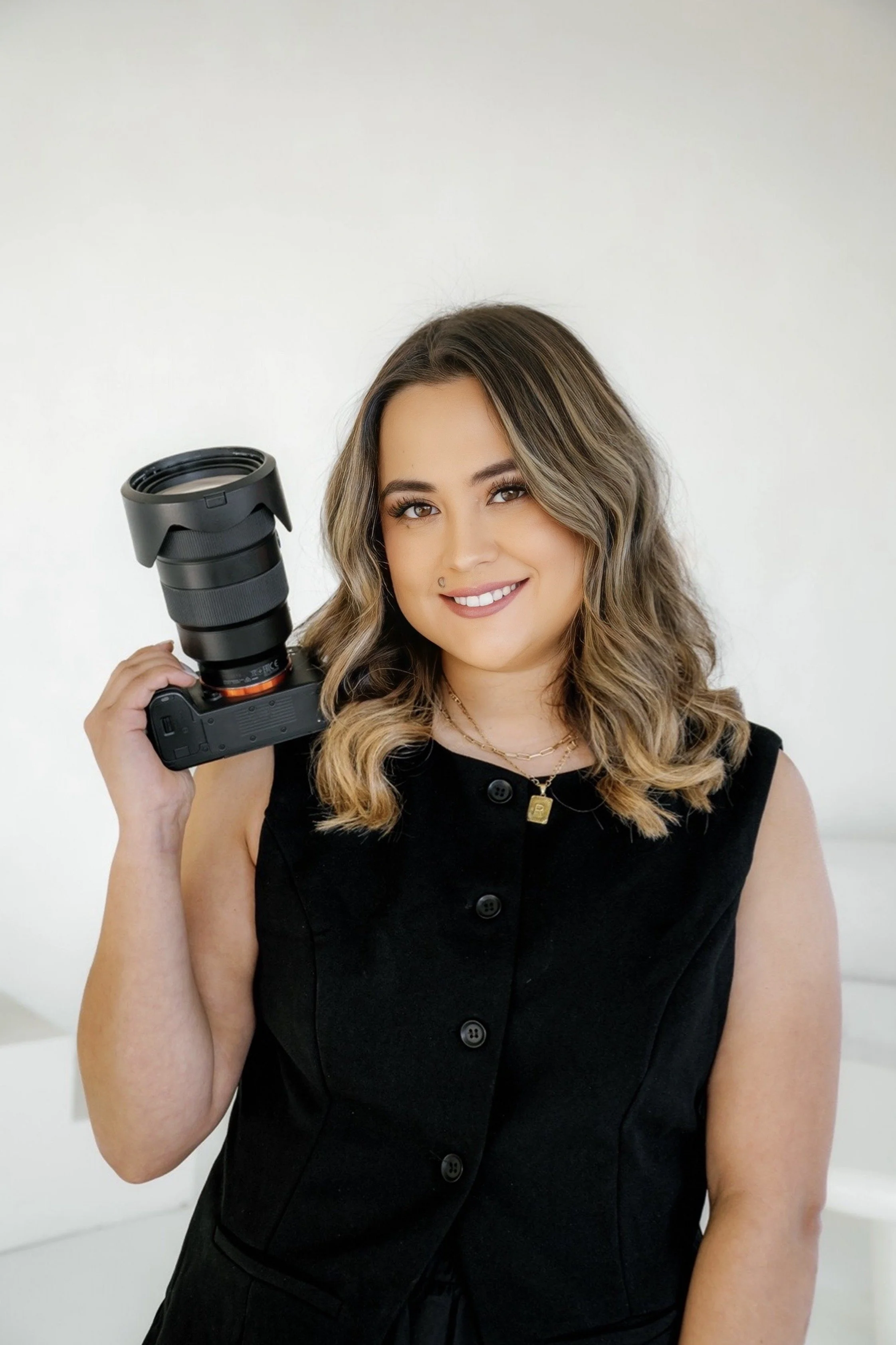 A woman with curly brown hair and glasses stands outdoors, smiling, while holding a tan straw hat with a wide brim. She is wearing a pinkish-red button-up shirt and blue jeans, leaning against a corrugated metal wall in a park-like setting with trees in the background.