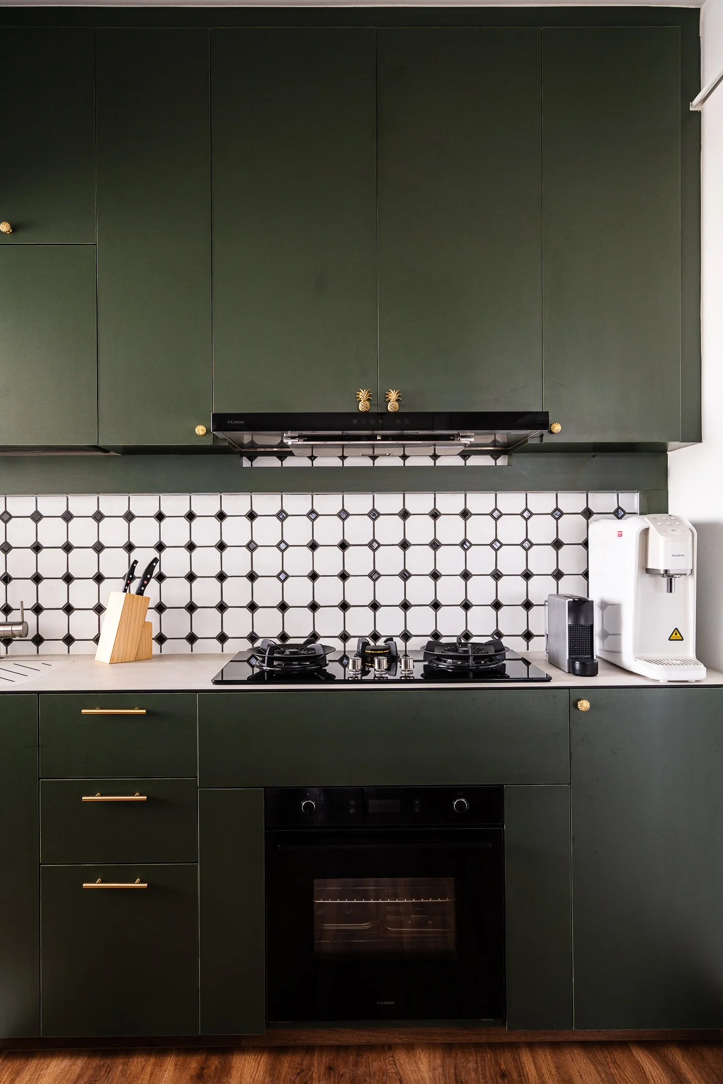 A vibrant forest green kitchen in a couple abode featuring unique gold pineapple-shaped cabinet handles and a geometric black-and-white tiled backsplash, showcasing bold interior design Singapore choices.