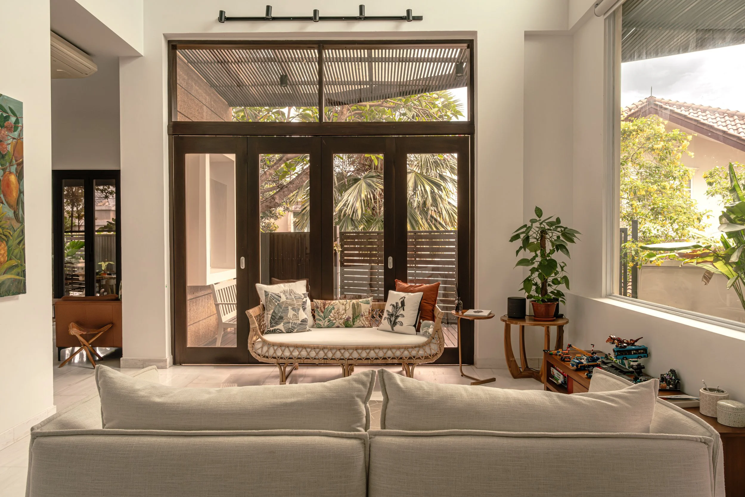 Tropical theme home interior design of living room of a 4 storey landed home at Bedok, with beige sofas, rattan bench with patterned cushions, large windows with a garden view, and indoor plants.