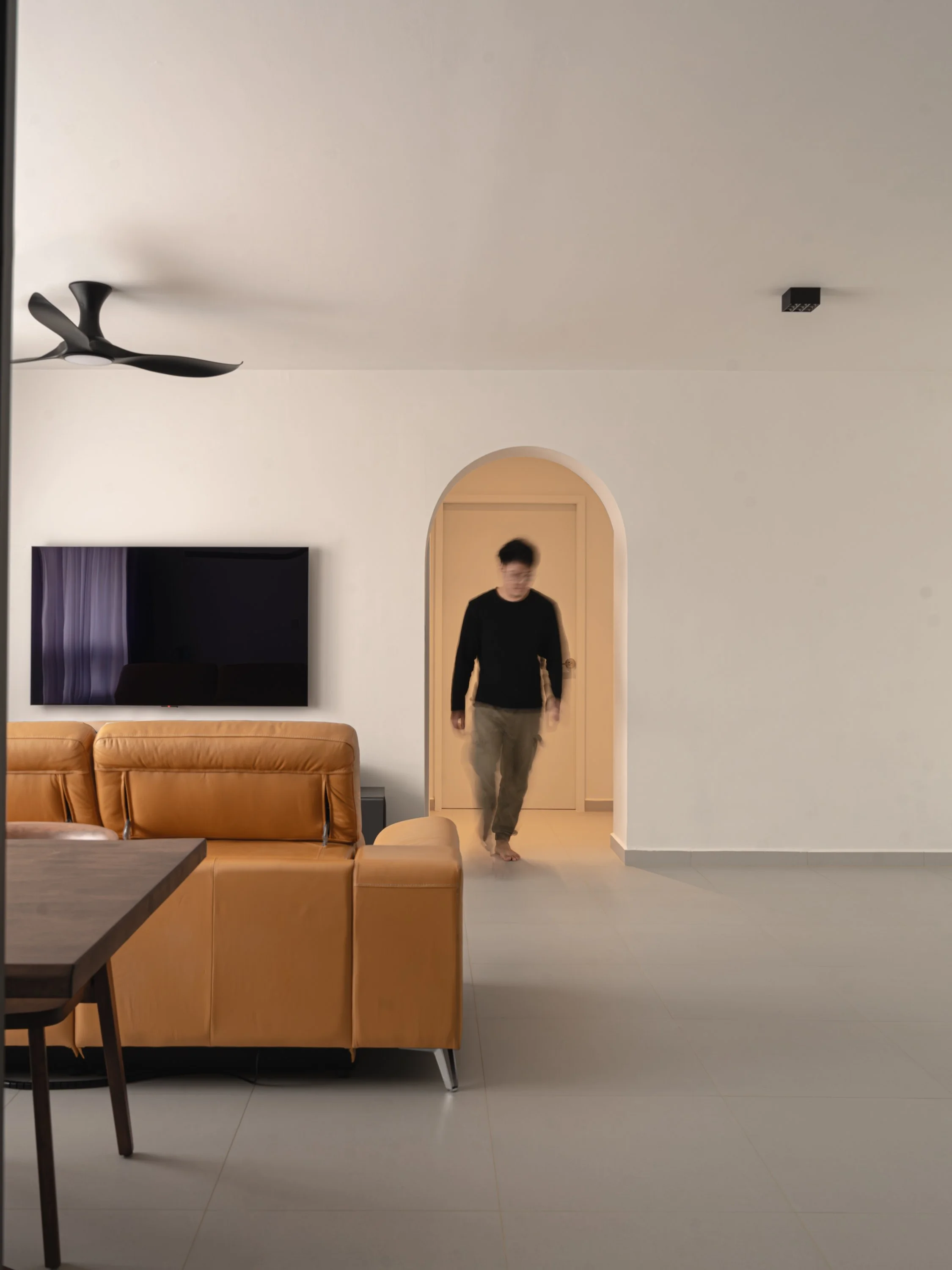 Modern living room with tan leather sofa, arched doorway, minimalist ceiling fan, and wall-mounted TV.
