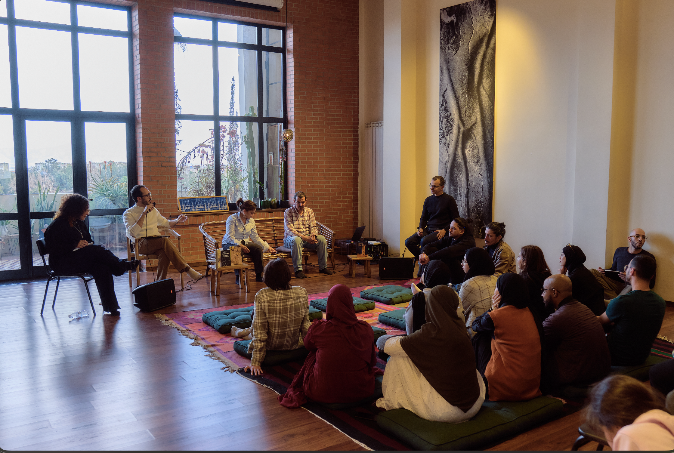 Un groupe de personnes participe à une discussion ou à un atelier dans une pièce avec de grandes fenêtres. Certains sont assis sur des coussins par terre, d'autres sur des chaises, face à un panel de intervenants.