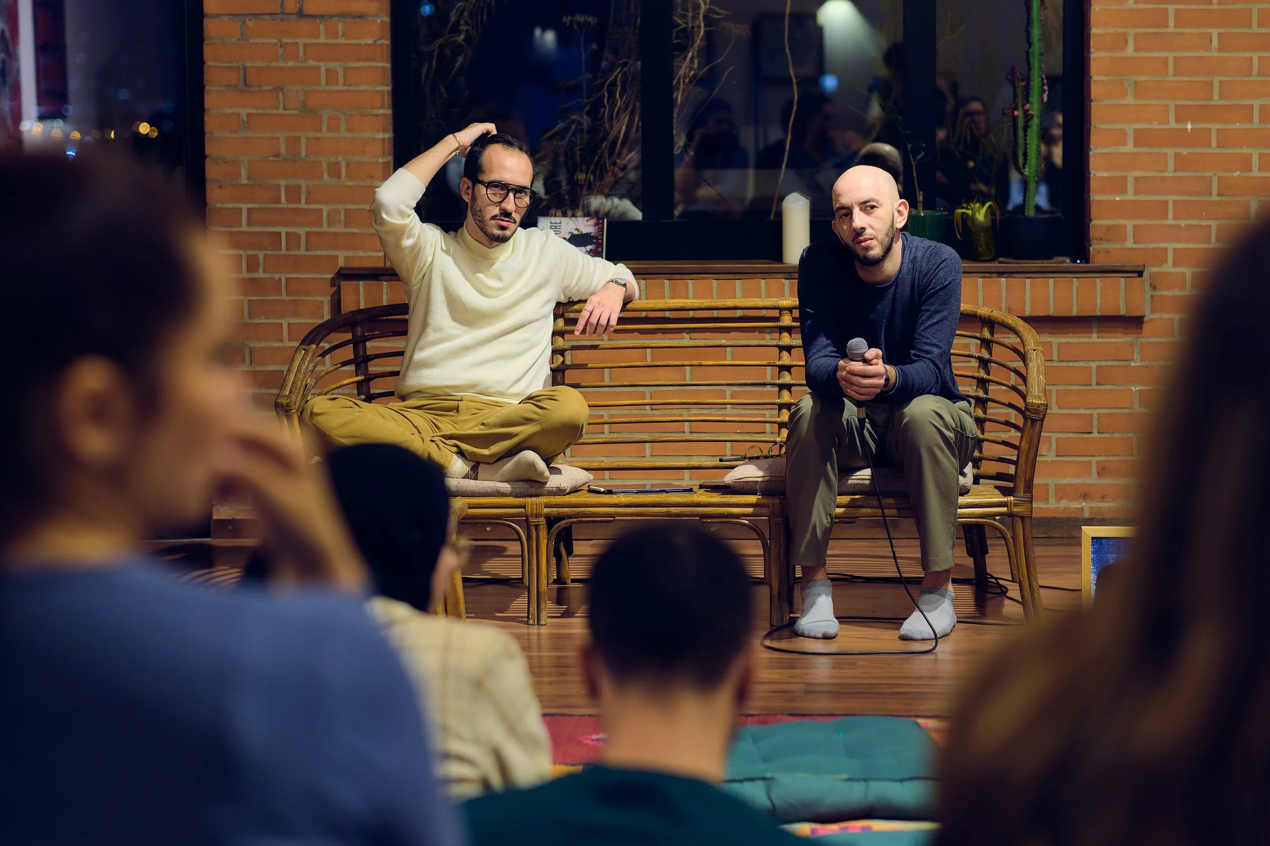 Deux hommes participent à une discussion ou un spectacle dans une salle avec un public. La scène est devant une fenêtre et un mur en briques, un homme en pull blanc et pantalon beige est assis en tailleur, l'autre en pull gris et pantalon vert est as