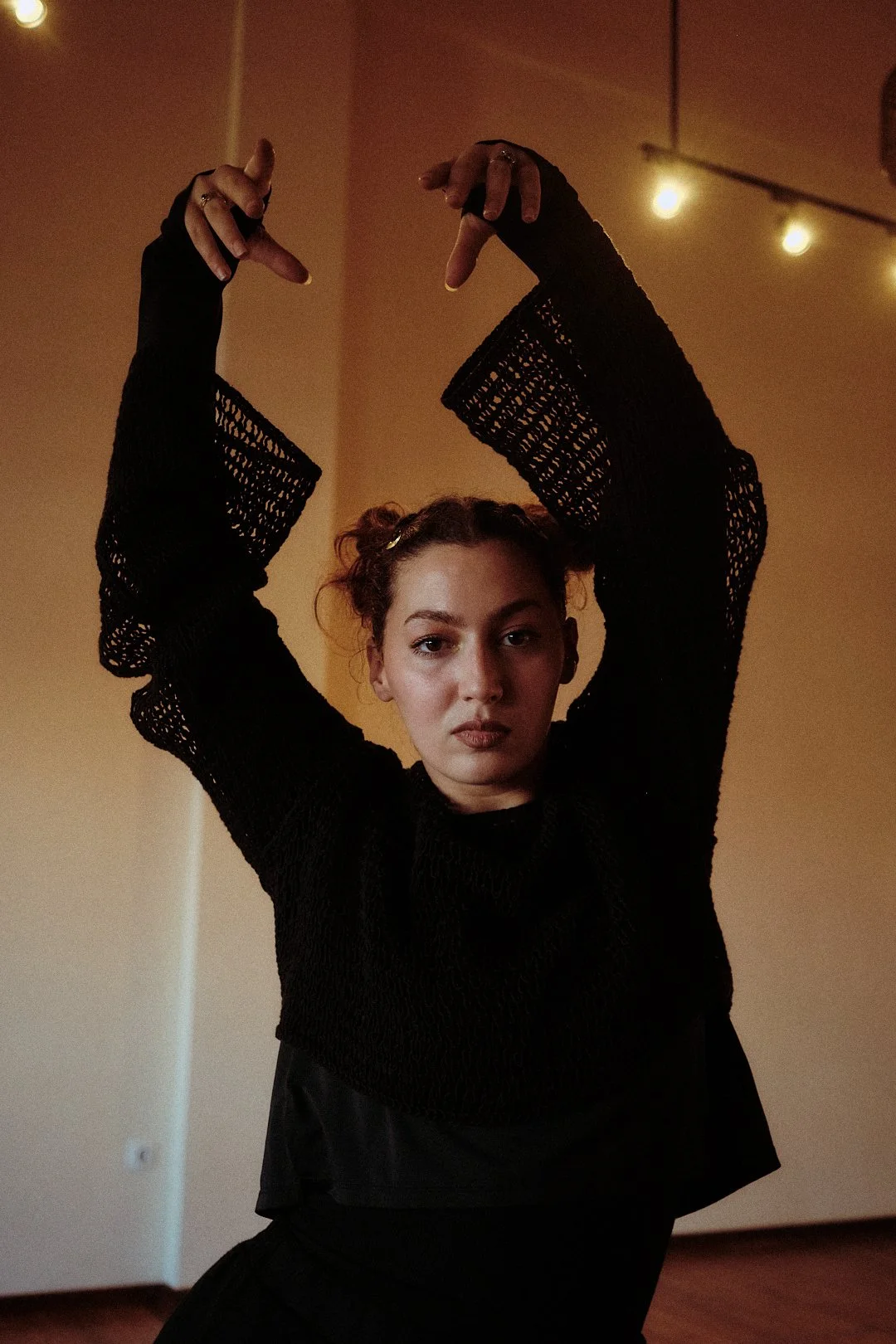 Femme portant une tenue noire, les bras levés, dans une pièce éclairée par une guirlande lumineuse.