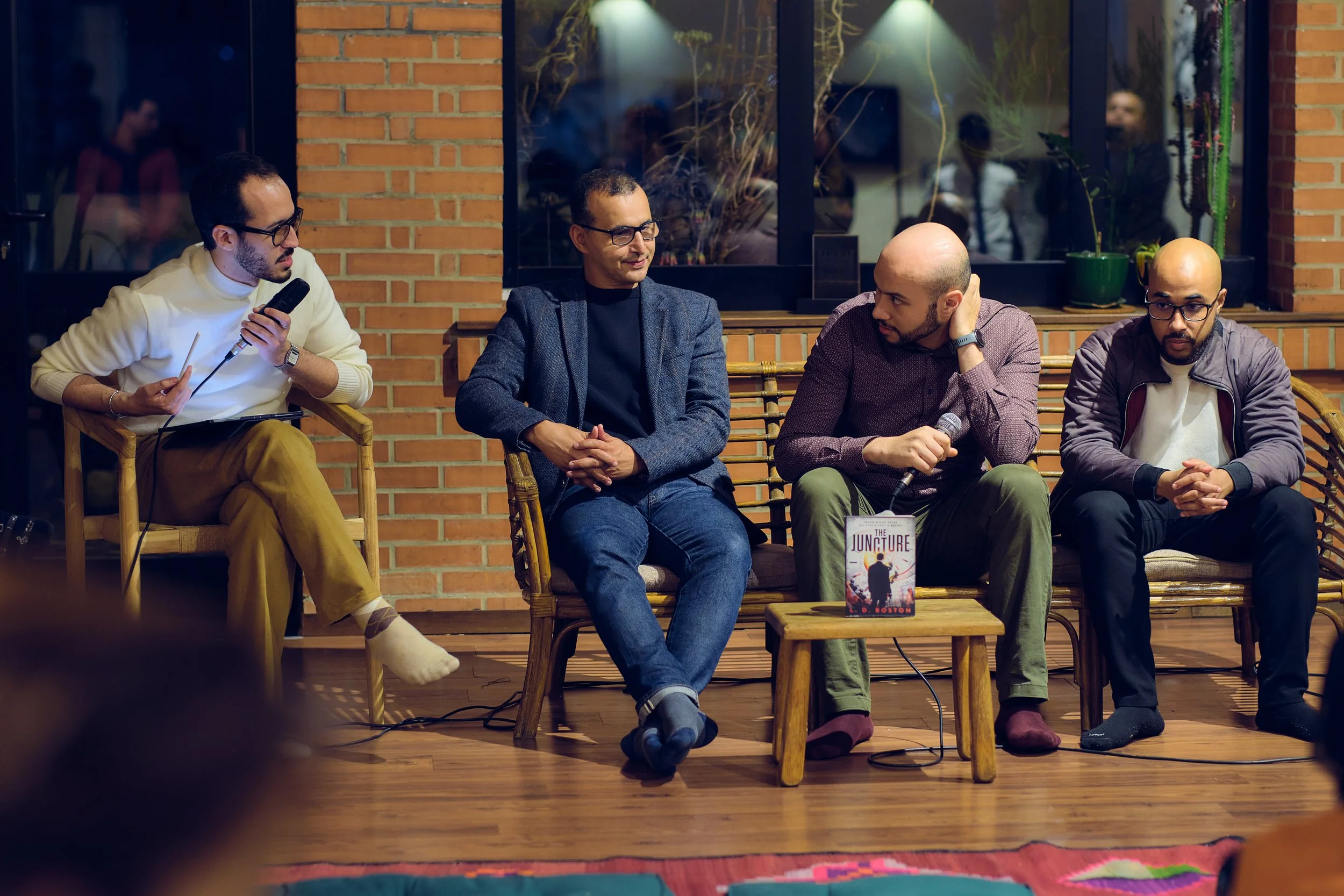 Quatre hommes assis sur un banc en bois lors d'une séance de discussion ou de présentation dans une salle avec mur en briques et grande fenêtre.