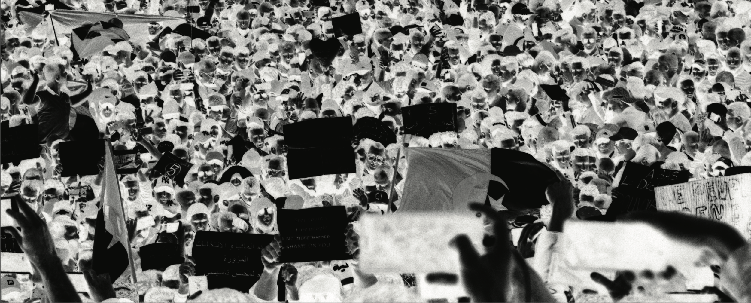 Une grande foule de personnes assistant à une manifestation, avec plusieurs drapeaux et panneaux, en noir et blanc.