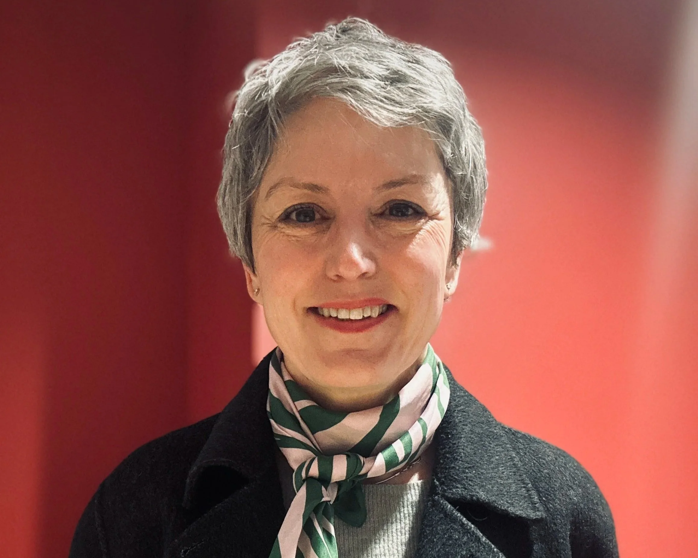 Close-up of a smiling  professional woman, Eline Strøm-Gundersen with short hair, wearing a grey suit jacket and a striped green and white silk scarf, standing against a red background.