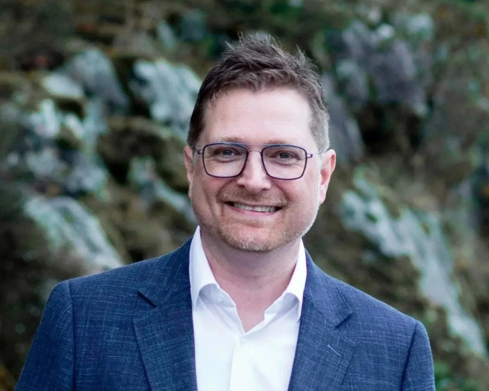 A man with short brown hair, glasses, and light facial hair, smiling and wearing a dark blue suit with a white shirt, standing outdoors with blurred green foliage in the background.