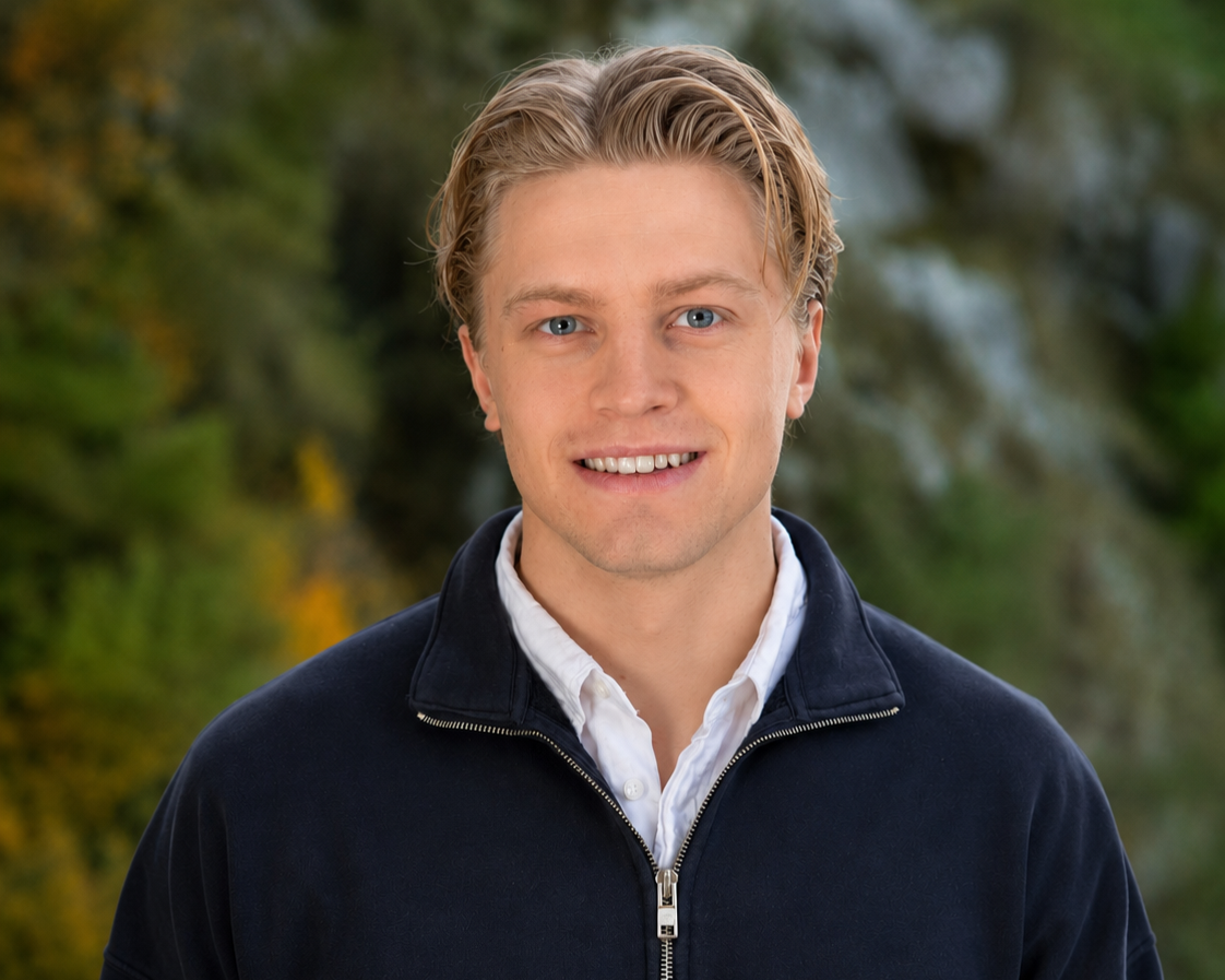Headshot of a young man with blonde hair, blue eyes, wearing a white collared shirt and a dark jacket, outdoors with blurred green foliage background.