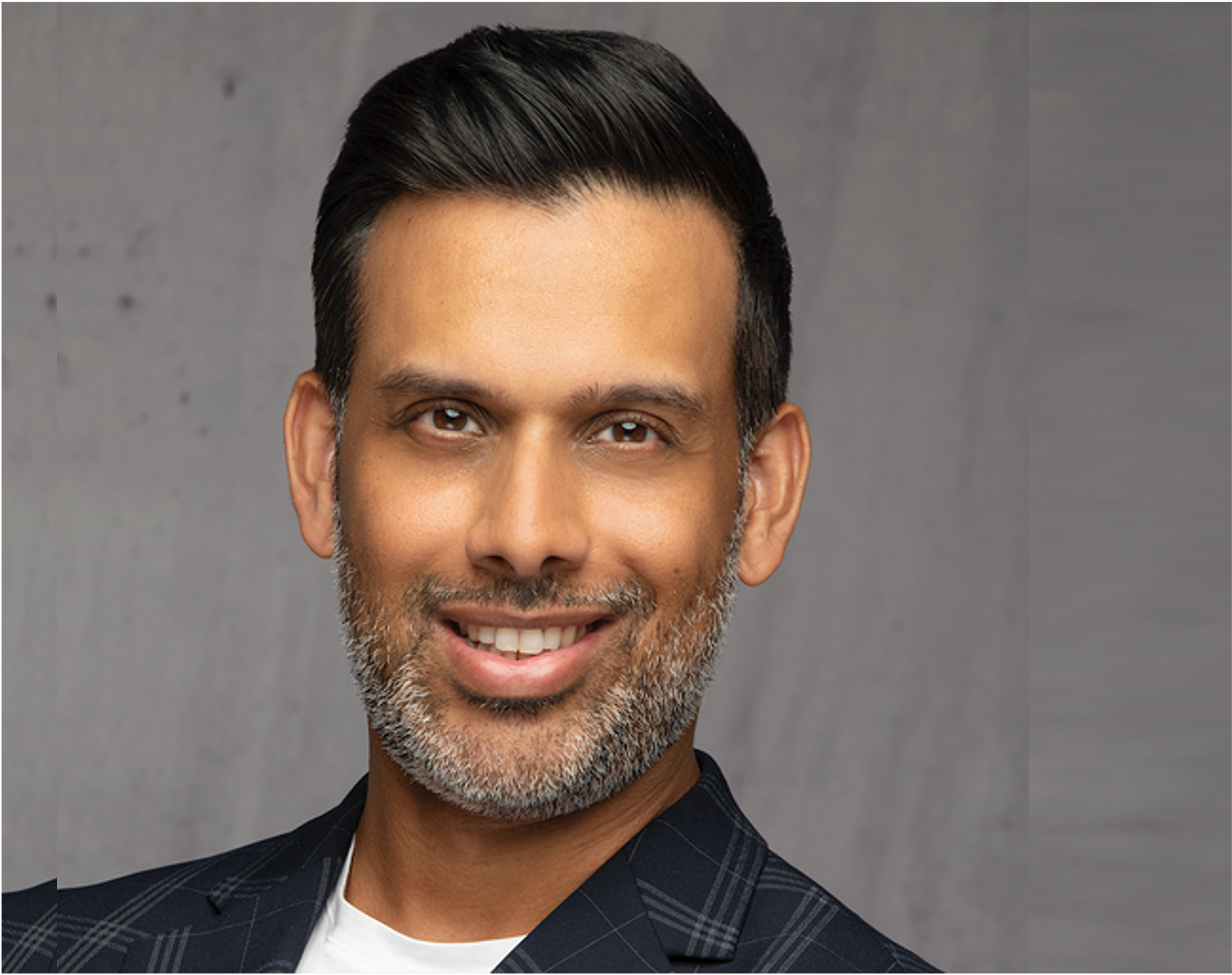A smiling middle-aged man, Ajay Khari,  with dark hair and a salt-and-pepper beard, wearing a dark plaid jacket over a white shirt, in front of a gray background.