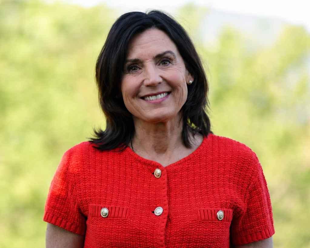 A woman with dark hair smiling outdoors, wearing a red textured short-sleeve top with gold buttons, against a blurred green and yellow background.