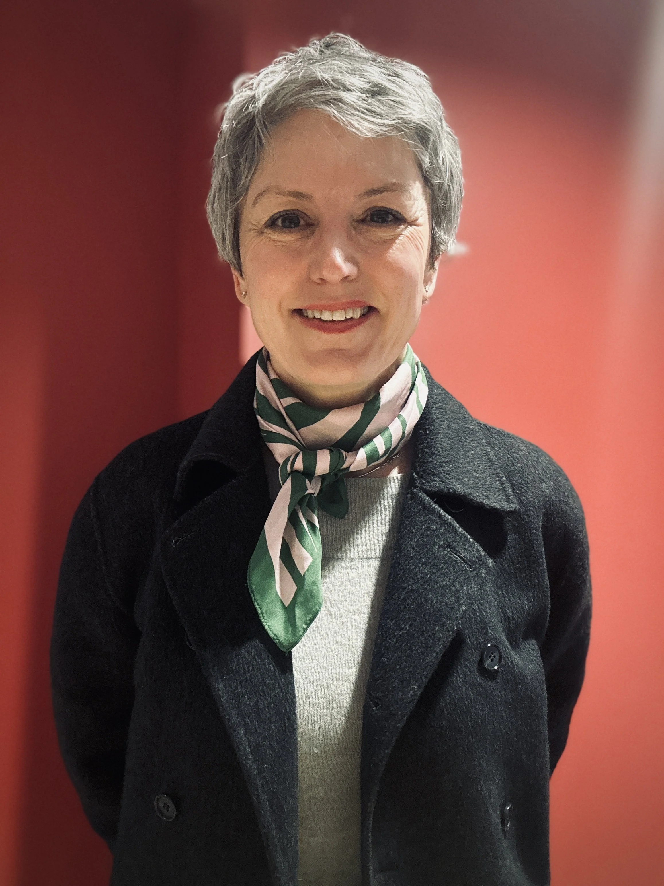 Smiling woman, Eline Strøm-Gundersen, with short gray hair wearing a dark coat, light gray sweater, and a green and white striped scarf, standing against a red background.