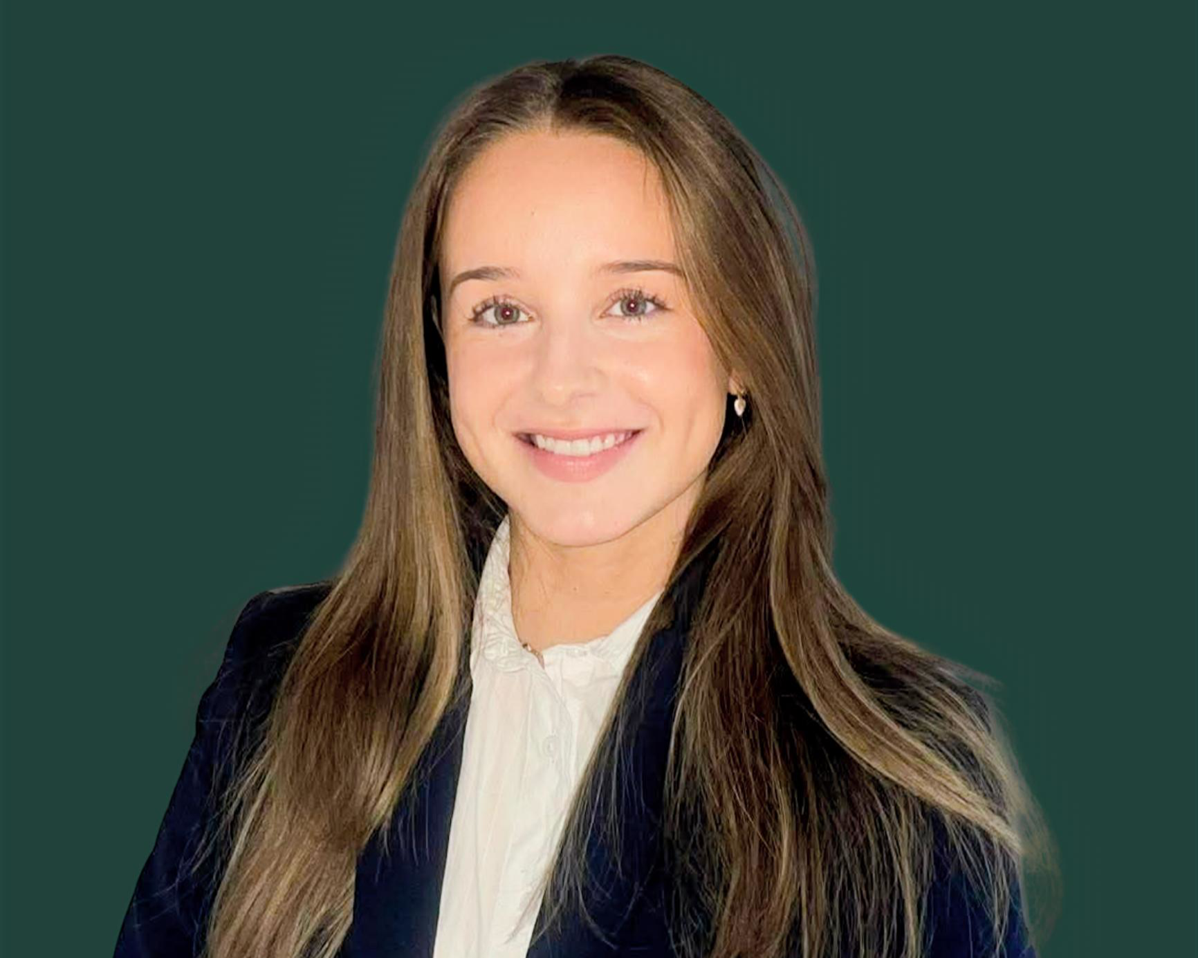 A young woman with long brown hair wearing a white blouse and a dark blazer, smiling at the camera, with a plain dark green background.