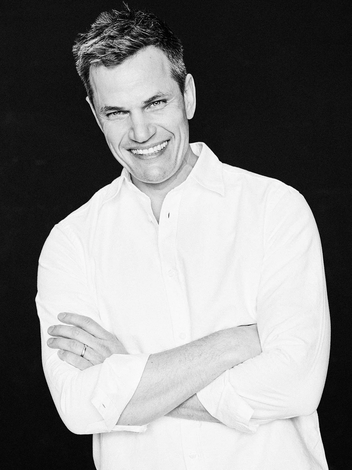 Schwarz-weiß Fotografie eines lachenden Mannes mit kurzen dunklen Haaren, in einem weißen Hemd mit aufgekrempelten Ärmeln, vor dunklem Hintergrund.