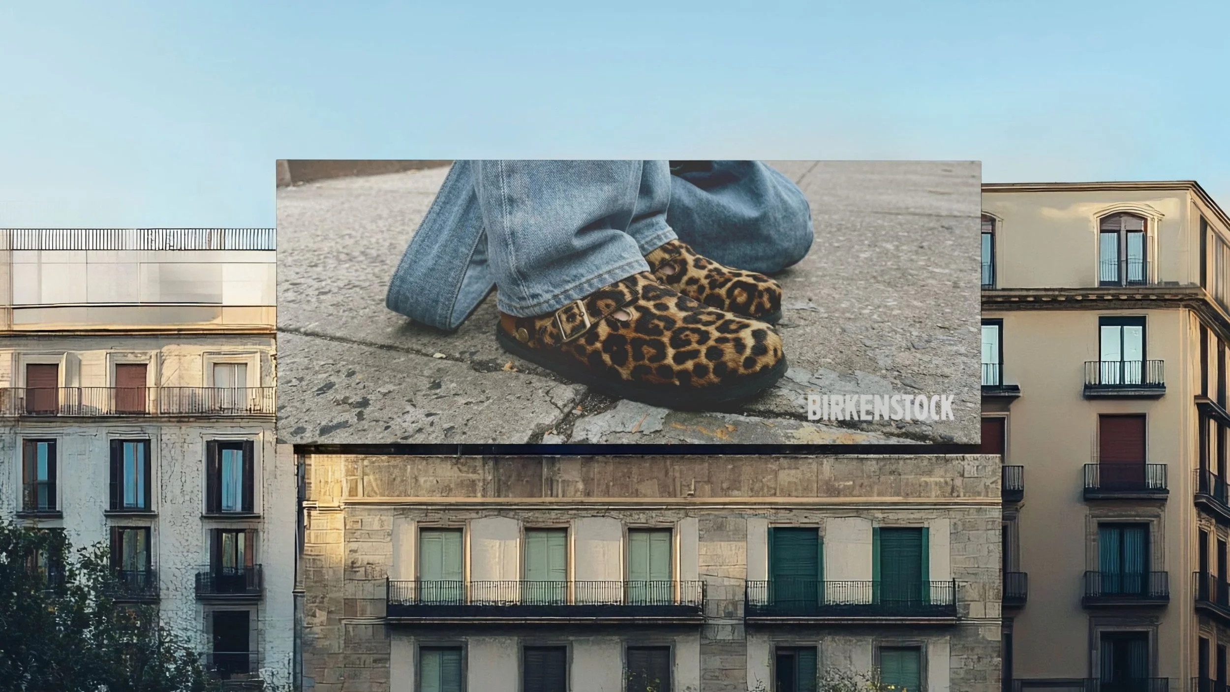 Straßenansicht mit mehreren alten Gebäuden, im Vordergrund ein Plakat mit einem Bild von bunten Leopardenfell-Schuhen. Das Plakat zeigt nur die Schuhe und die Jeans der Person, keine weiteren Details. Der Himmel ist bläulich. Auf dem Plakat steht das Wort "BIRKENSTOCK".
