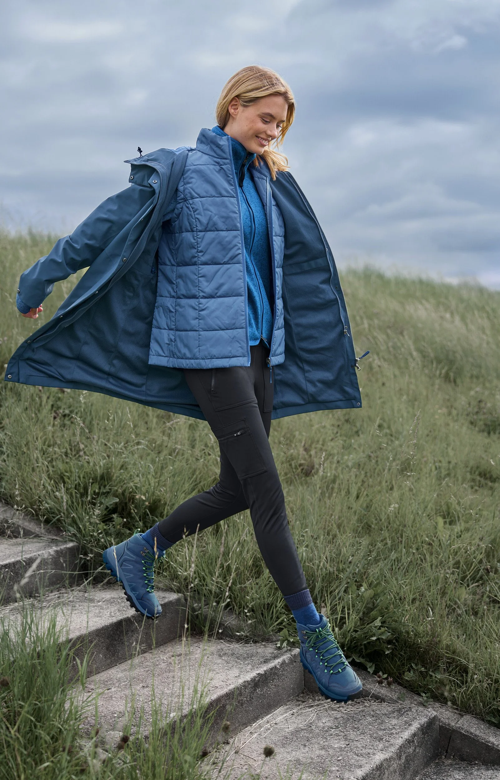 Frau beim Wandern auf Treppen im Freien bei bewölktem Himmel, trägt blaue Outdoor-Kleidung und Wanderschuhe.