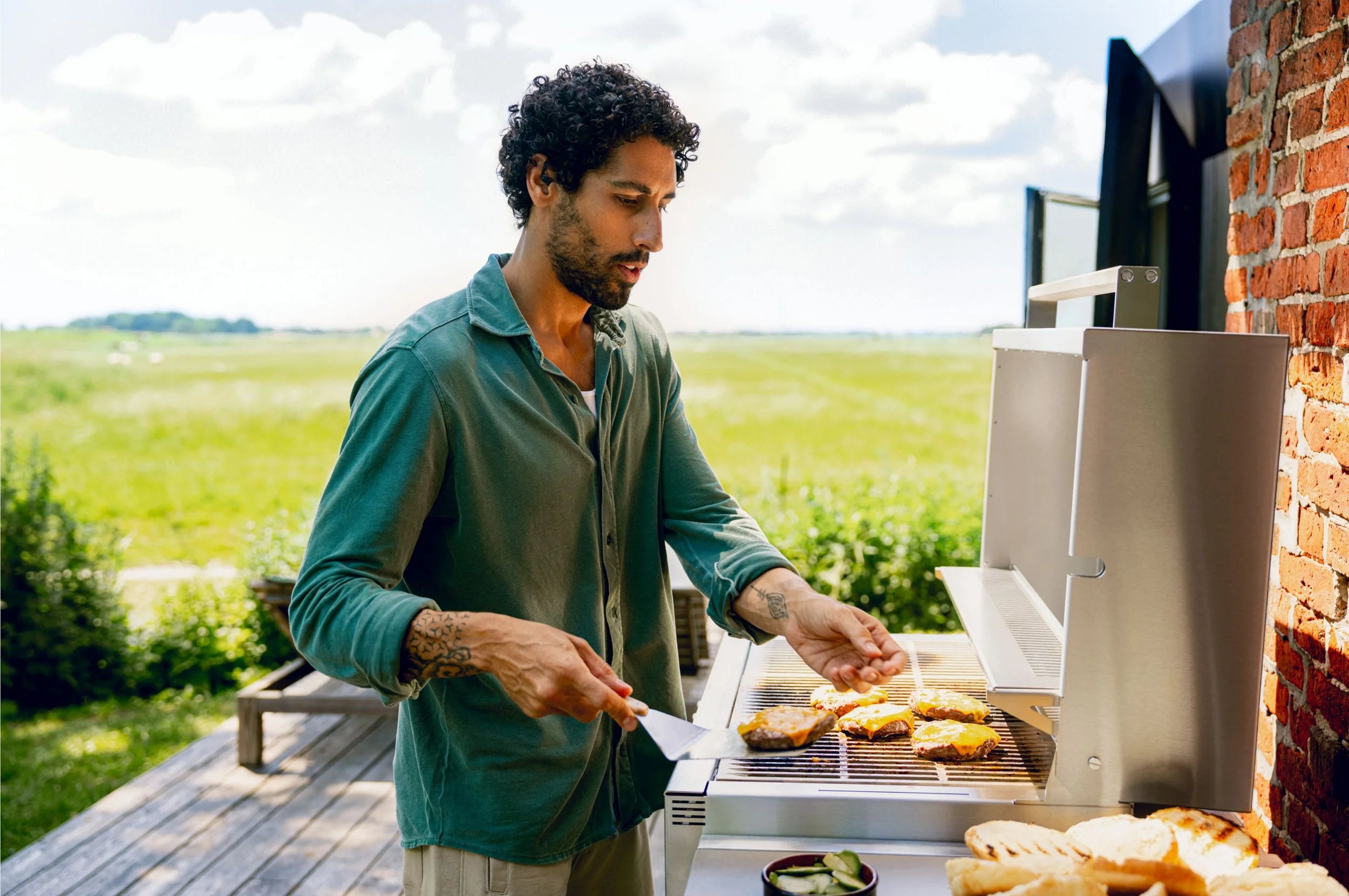 Mann grillt Fleisch und Gemüse im Freien auf einem Grill mit grünem Landschaftshintergrund.