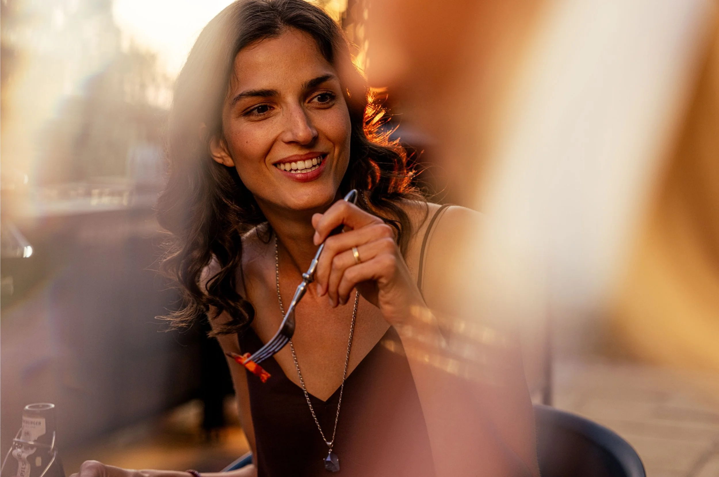 Eine lachende junge Frau mit braunen, lockigen Haaren, die einen Löffel hält, bei Sonnenuntergang draußen.