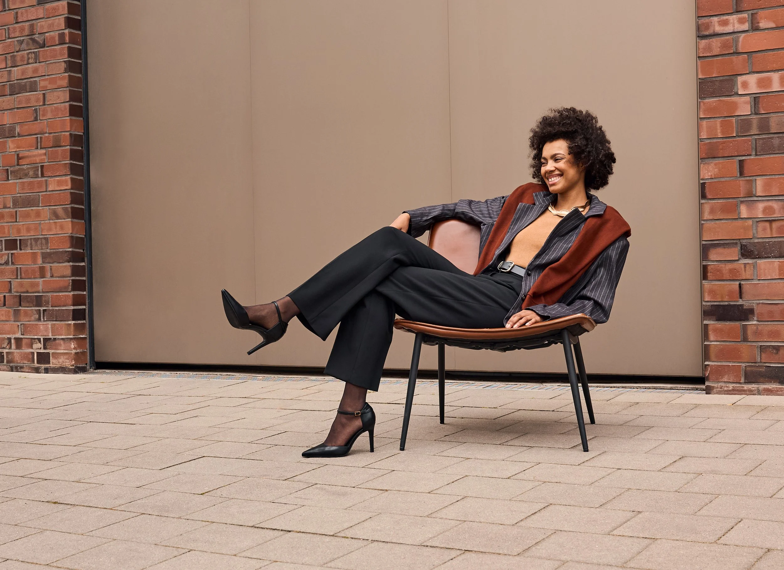 Eine Frau sitzt auf einem Stuhl vor einer Wand, lächelt und trägt elegante Kleidung mit hohen Schuhen.