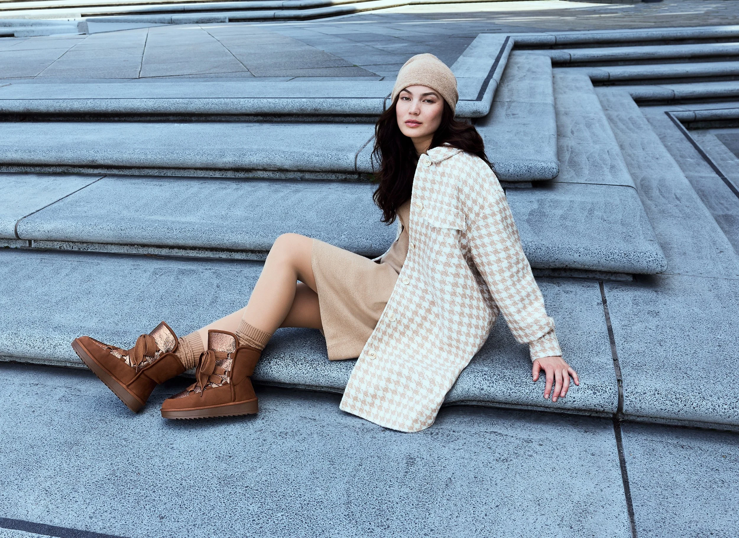 Eine junge Frau sitzt auf einer Treppe aus grauem Stein, trägt einen beigen, karierten Mantel, eine beige Pumphose, braune Boots, ein beigefarbenes Hut und schaut in die Kamera.
