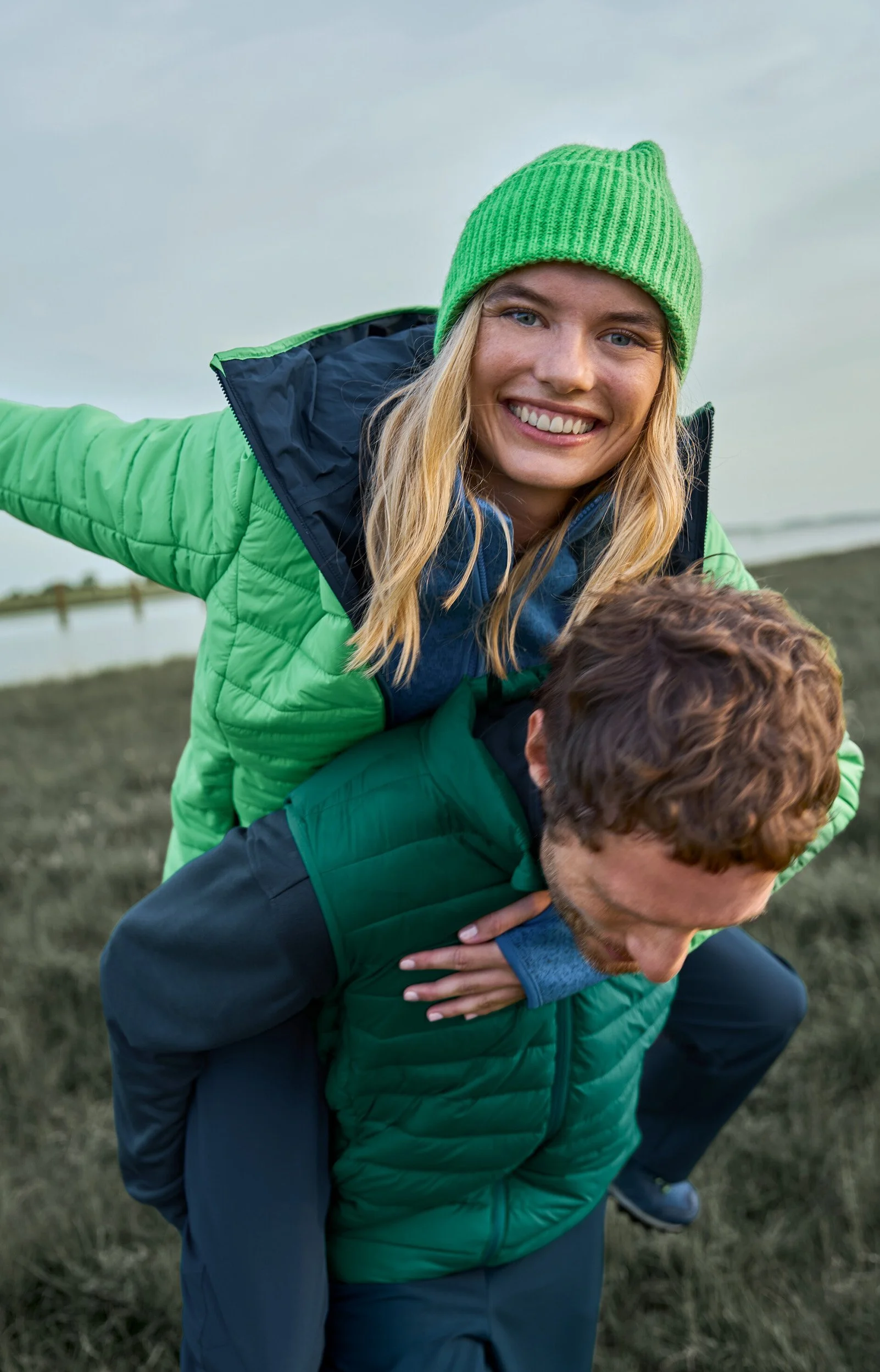 Ein lächelnder Frau trägt einen Jungen auf dem Rücken in einer Outdoor-Umgebung an einem bewölkten Tag, beide tragen grüne Jacken.