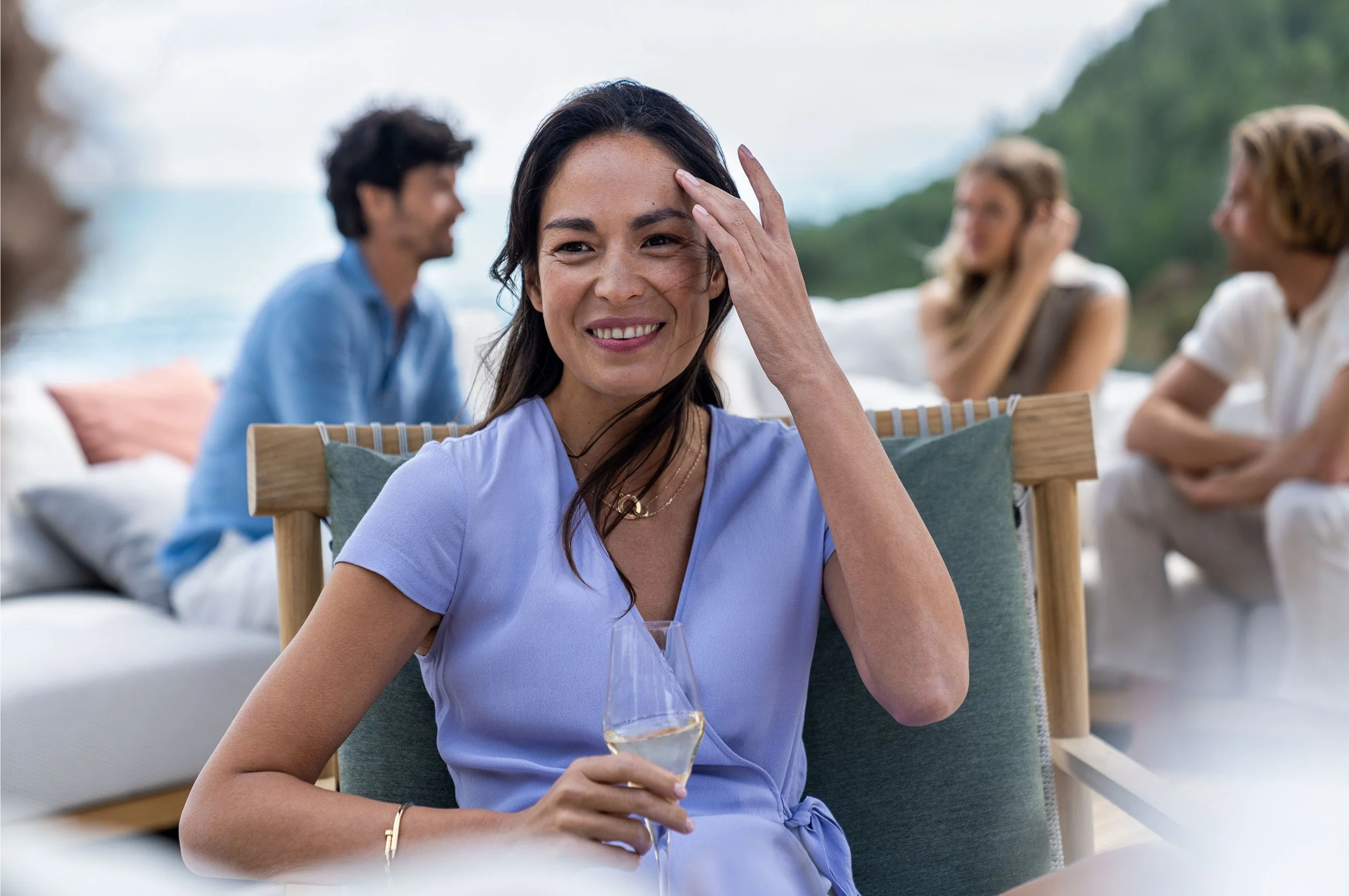 Eine Frau sitzt draußen mit einem Glas Sekt, lächelt und berührt ihren Kopf, im Hintergrund sind drei weitere Menschen im Gespräch bei einem Treffen im Freien, mit Blick auf das Wasser und grüne Hügel.