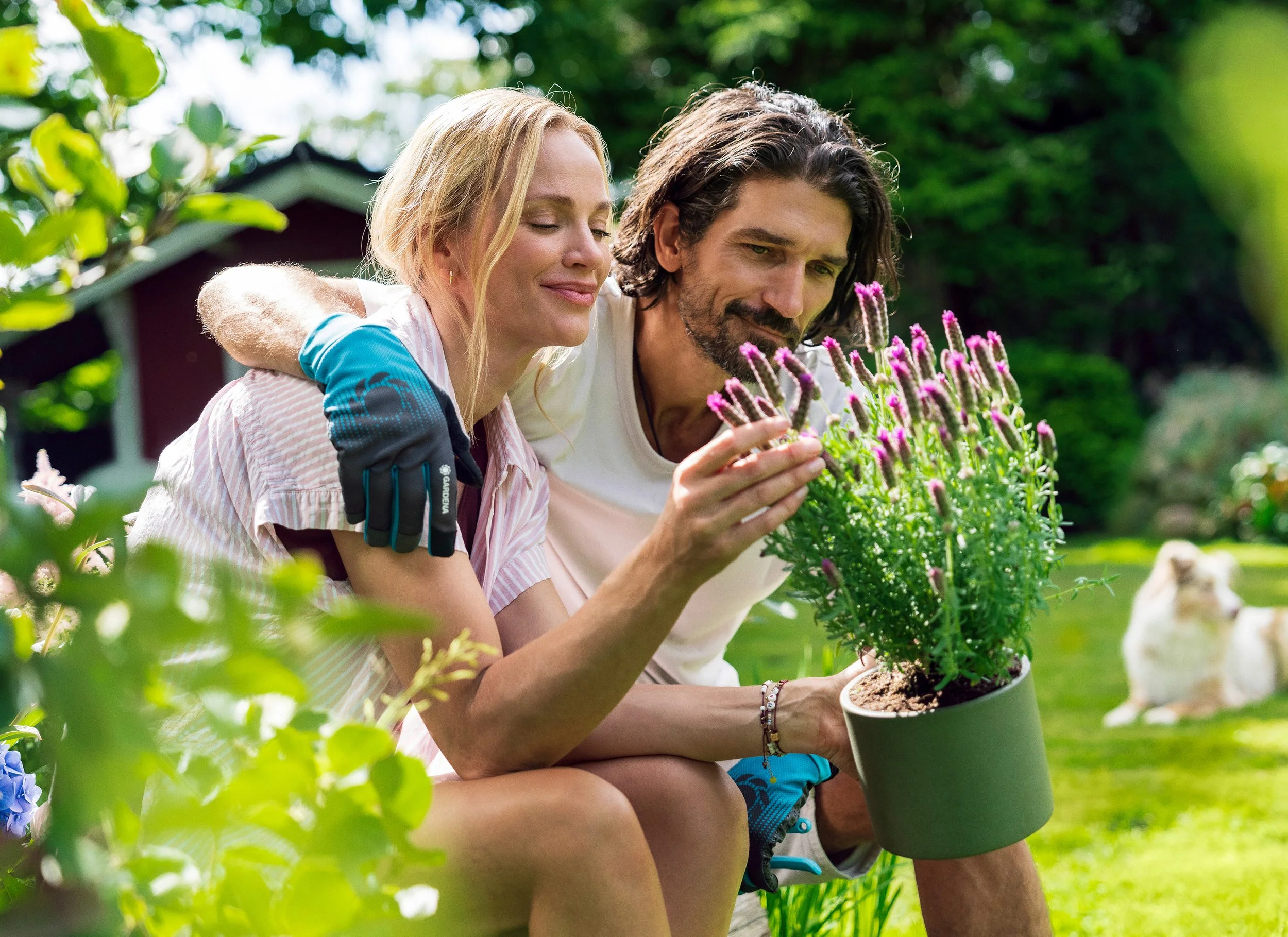Ein lachendes Paar, das im Garten sitzt, während die Frau eine Topfpflanze mit pinken Blumen hält.
