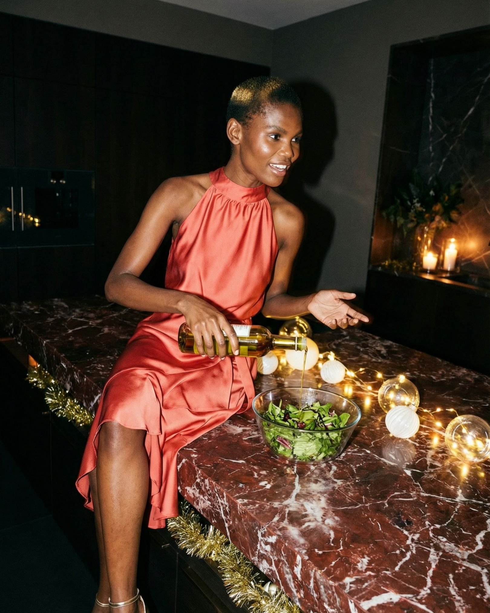 Eine Frau in einem roten Kleid macht einen Salat in einer modernen Küche, umgeben von weihnachtlicher Dekoration mit Lichterketten und Kerzen.