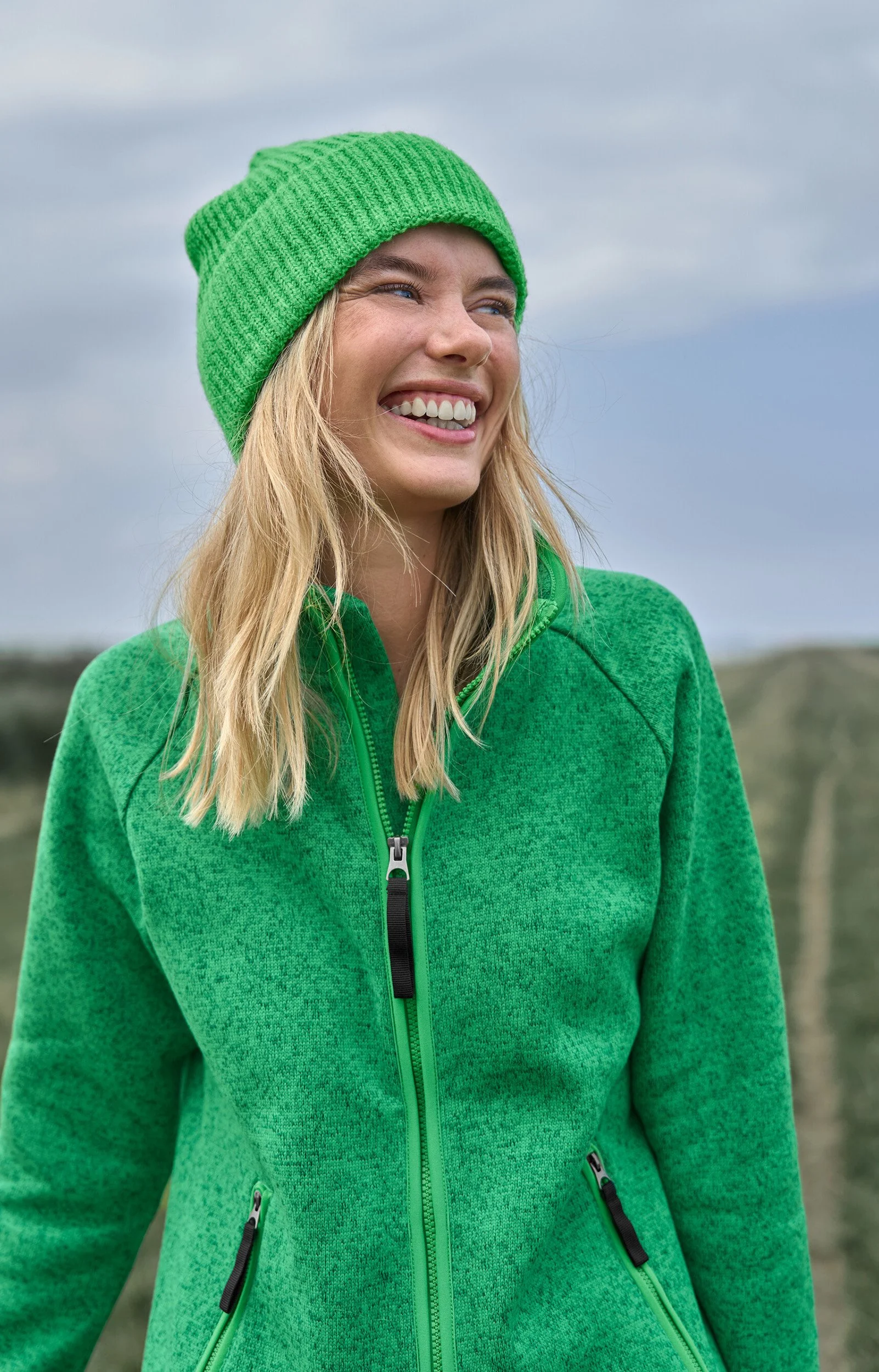Lächelnde junge Frau in grüner Jacke und grüner Mütze im Freien bei bewölktem Himmel.