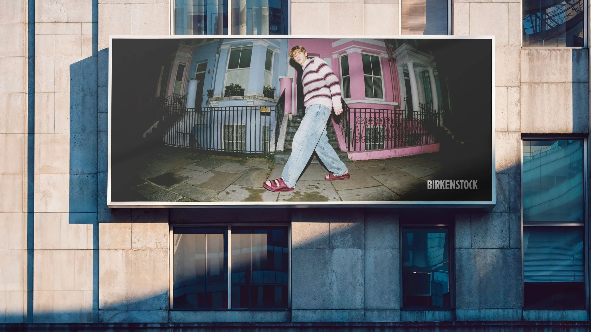 Ein Plakat an der Fassade eines Gebäudes zeigt einen jungen Menschen, der bei Nacht vor einem bunter Haus steht, mit dem Text 'BIRKENSTOCK' in der Ecke.