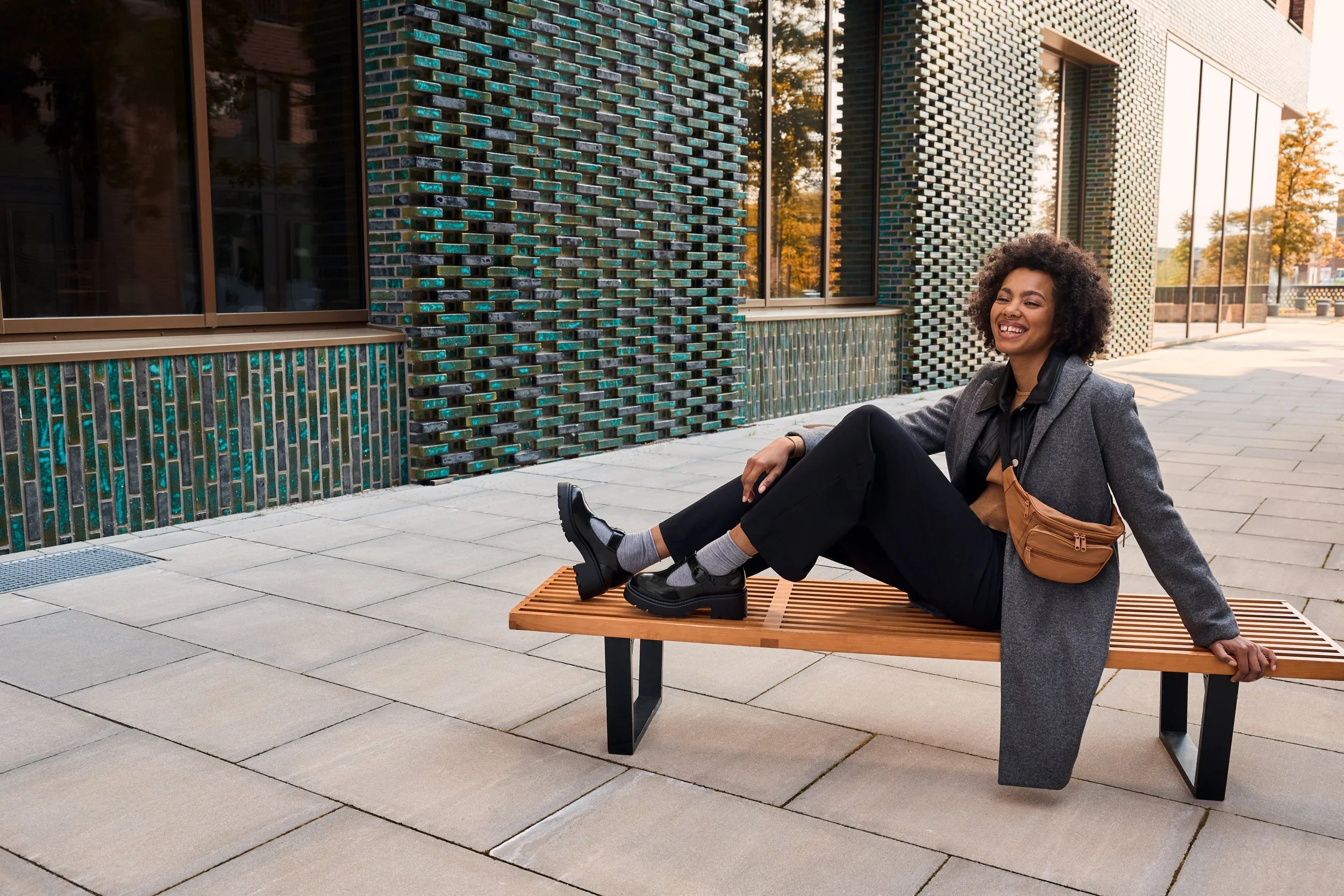 Junge Frau mit dunklen Locken sitzt lächelnd auf einer Holzbank vor einem modernen Gebäude mit bunten Fliesenfassaden, trägt schwarze Schuhe, grauen Mantel und braune Tasche.