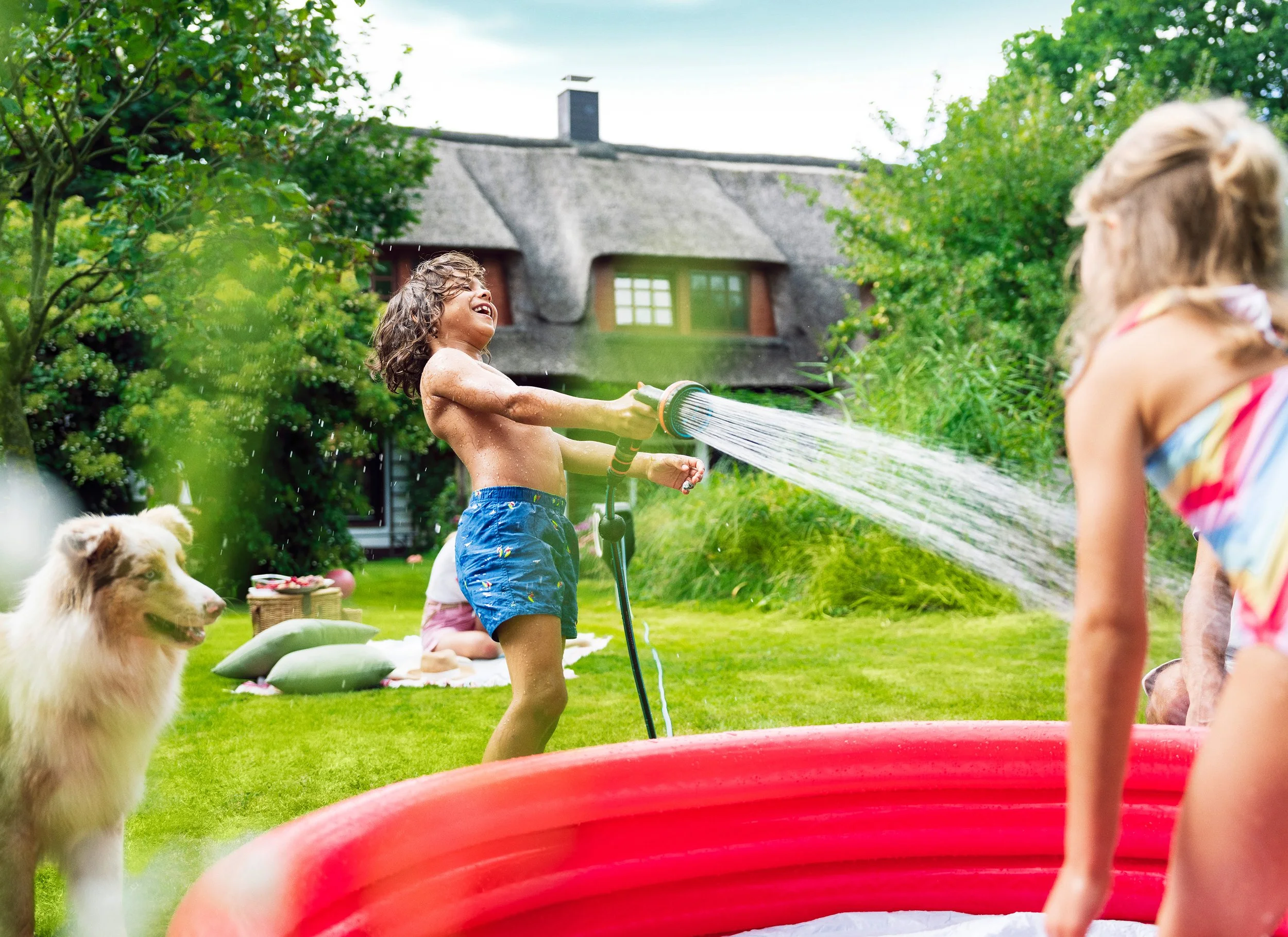 Kinder spielen mit Wasser im Garten, eine Gruppe badet und lacht, während Wasser mit einem Gartenschlauch sprüht. Im Hintergrund ein Hund und eine Familie, die sich entspannt auf einer Decke sonnt.