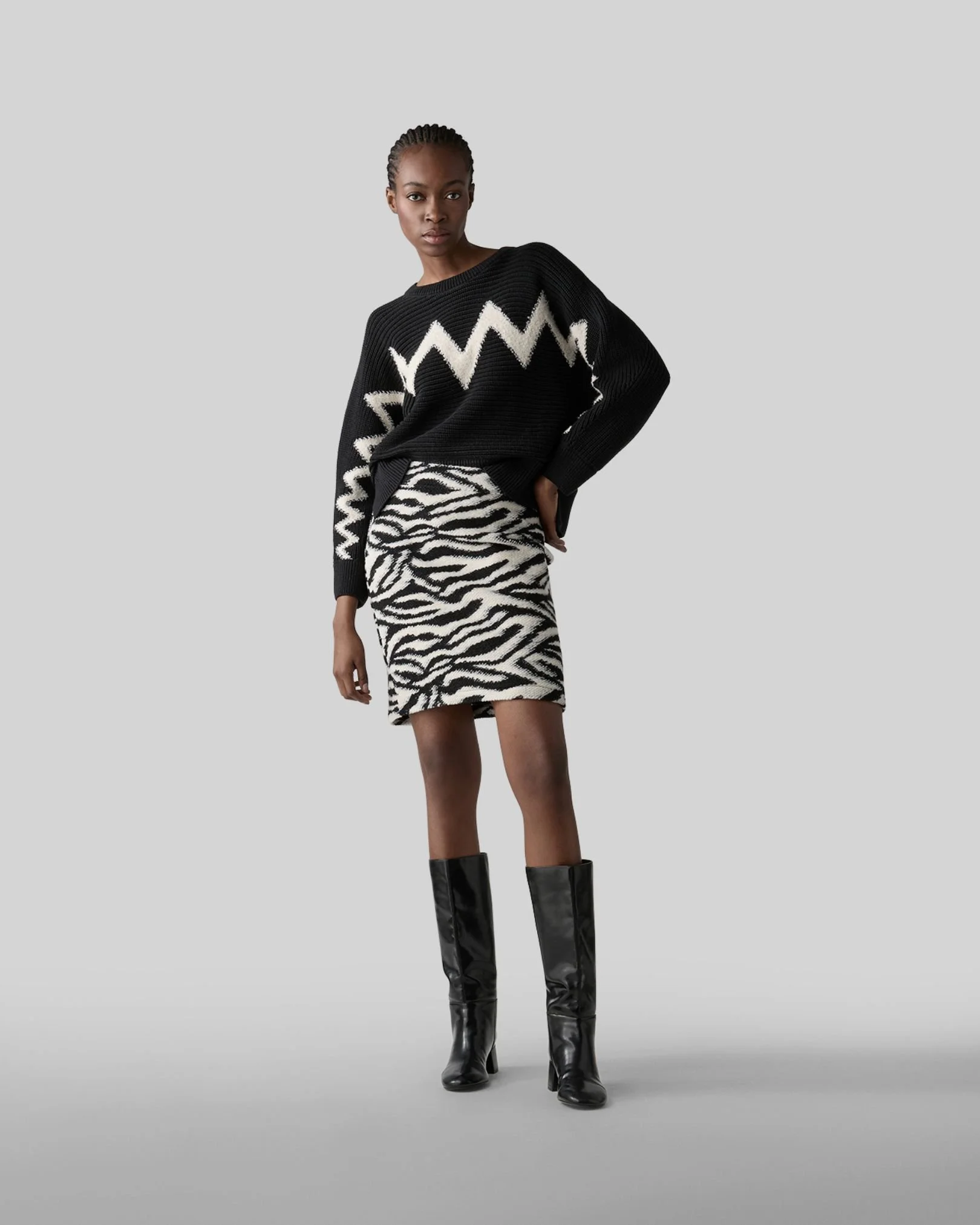 Eine Frau trägt einen schwarzen Pullover mit weißen Zickzack-Mustern, eine kurze schwarzen und weißen Zebra-Muster-Rock und hohe schwarze Stiefel, vor einem grauen Hintergrund.