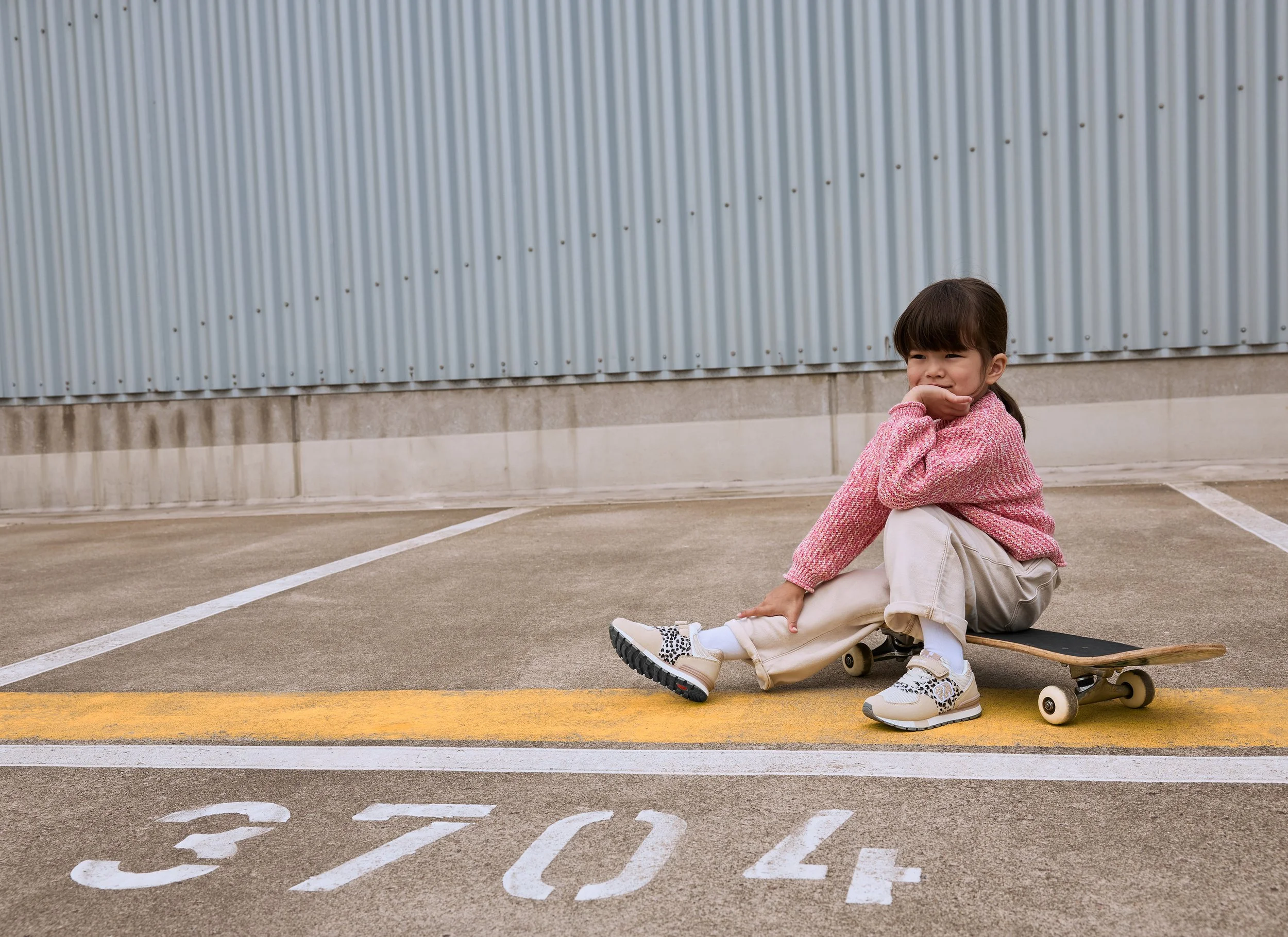 Ein Mädchen sitzt auf einem Skateboard auf einem Parkplatz mit weißen Nummern und gelben Linien vor einer Metallwand.
