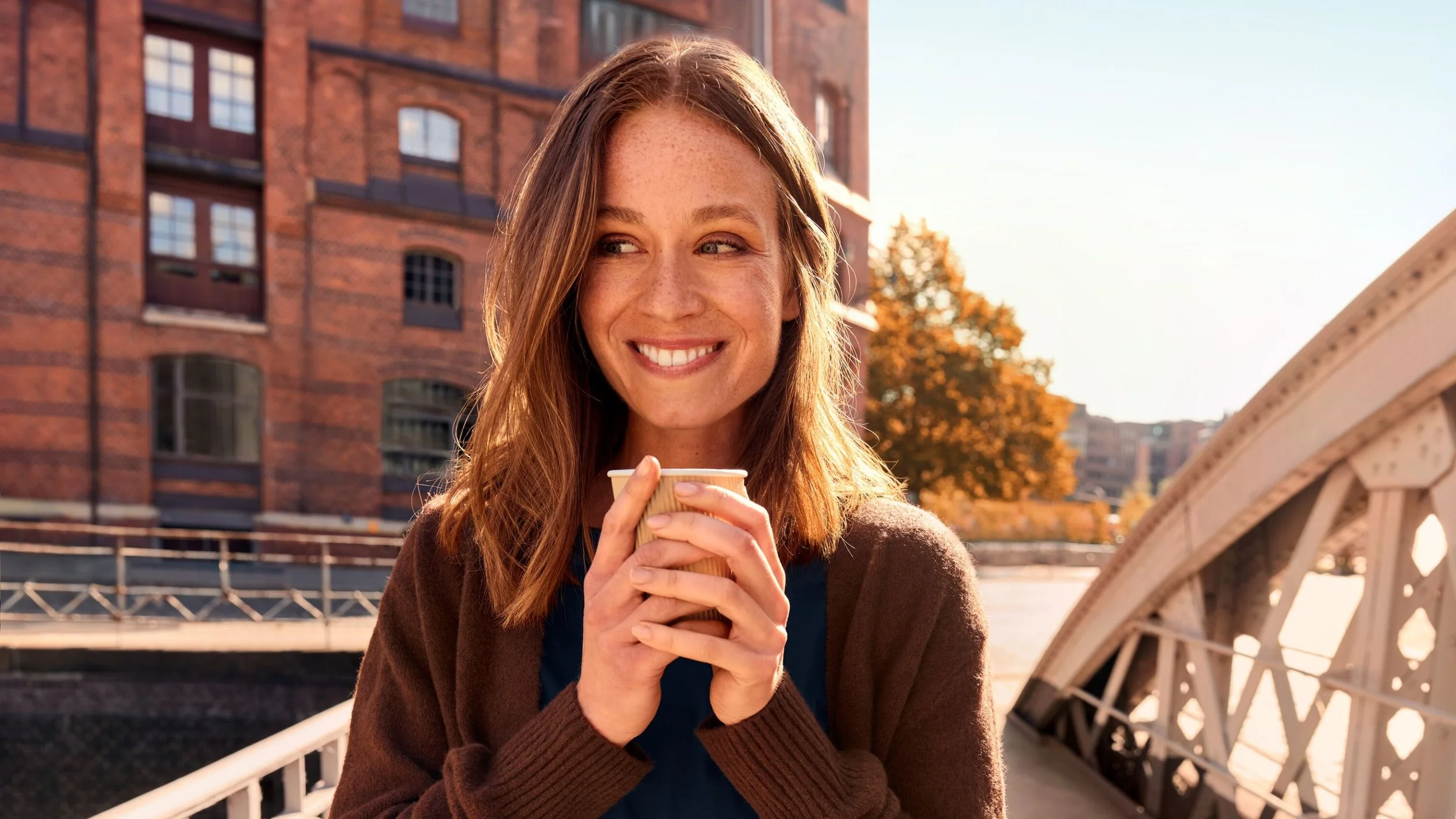 Junge Frau mit braunen Haaren, lächelnd, hält eine Heißgetränk in einer Papp-Mehrweg-Tasse auf einer Brücke bei Sonnenuntergang, Stadt im Hintergrund.