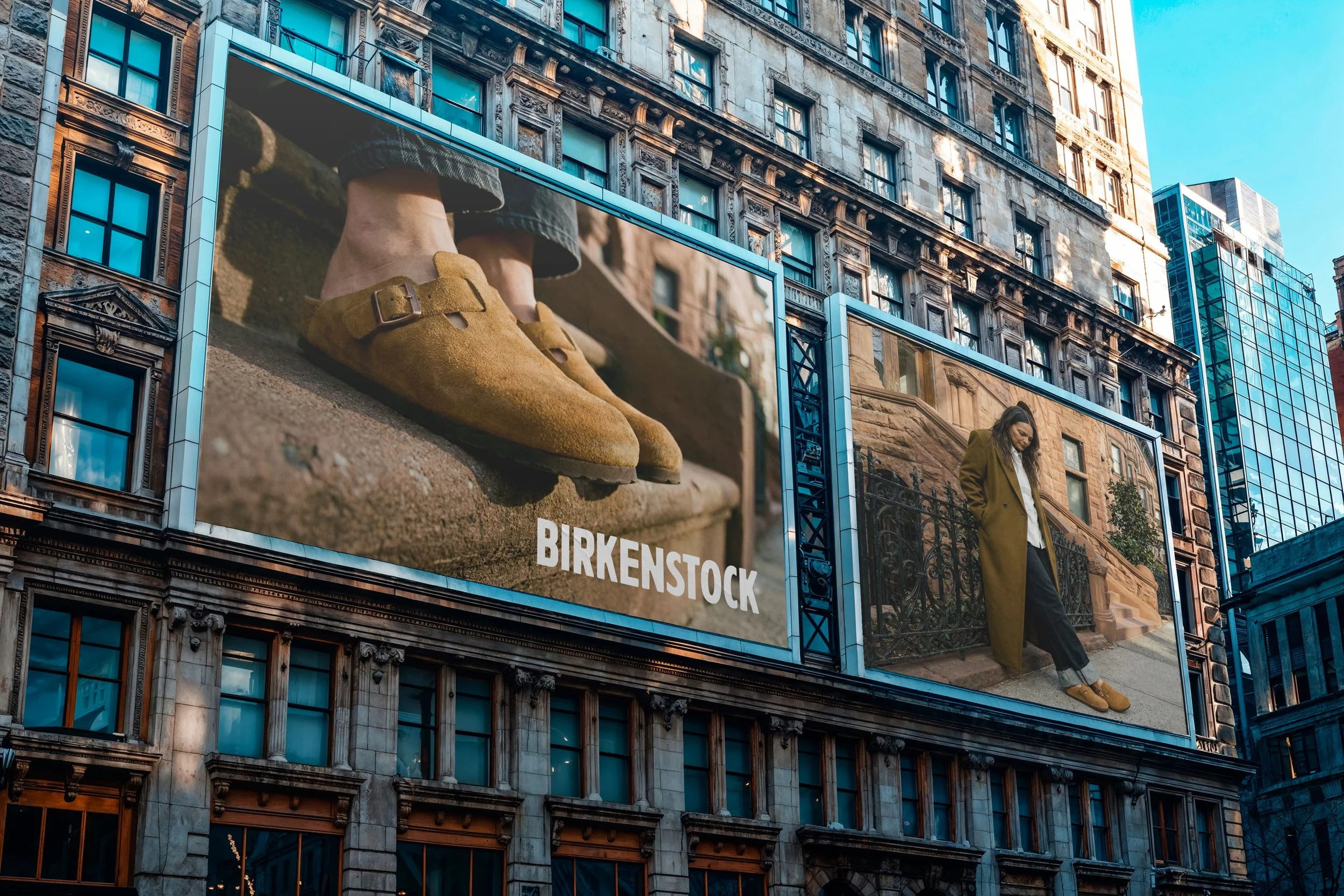 Große Werbetafeln an einem Gebäudekomplex in einer Stadt zeigen Schuhe und eine Frau in einem Mantel. Das Unternehmen BIRKENSTOCK ist sichtbar.