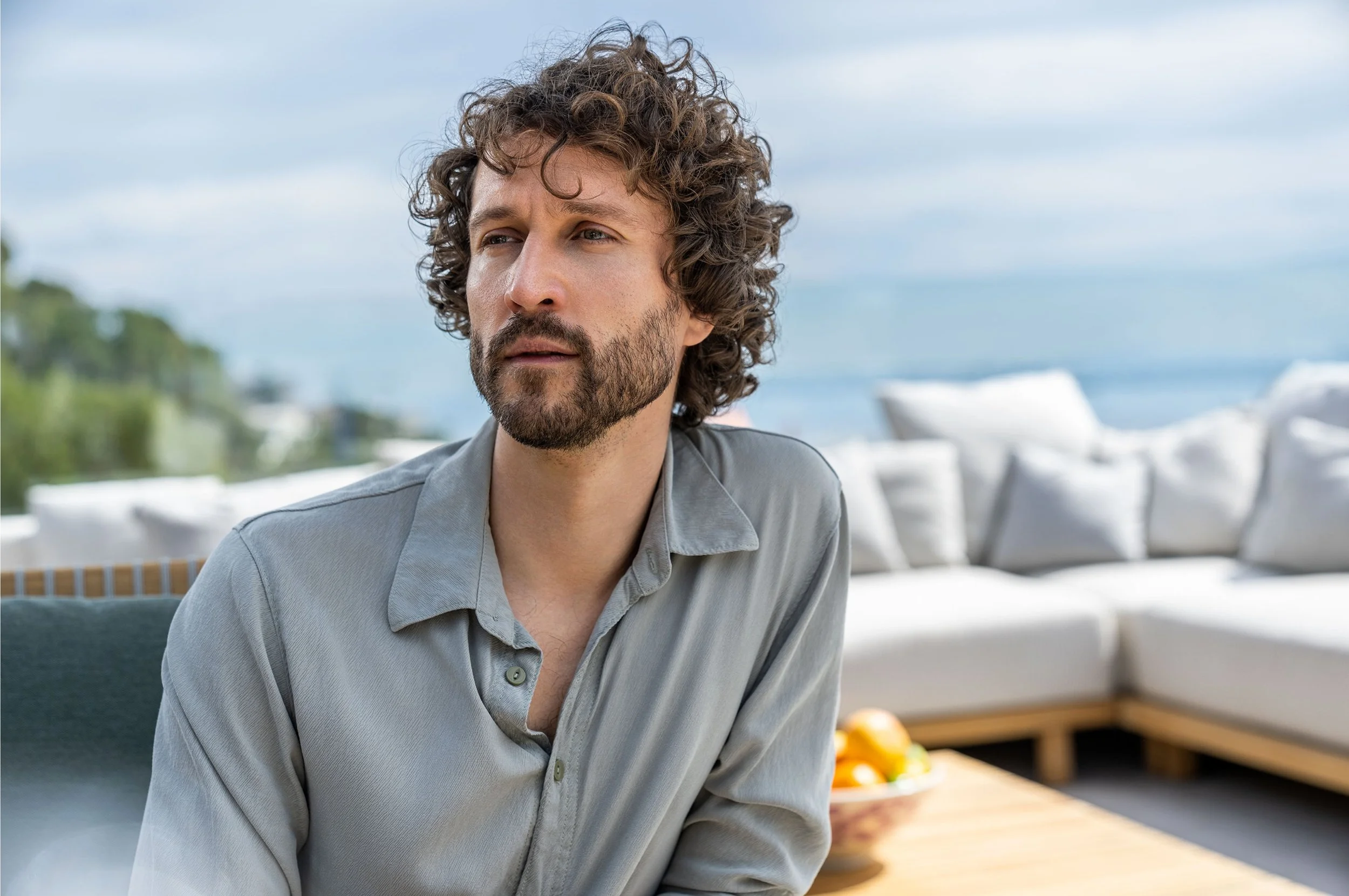 Ein Mann mit lockigem braunem Haar und Bart sitzt im Freien auf einer Terrasse mit Blick auf das Meer, im Hintergrund ein Sofa mit weißen Kissen.