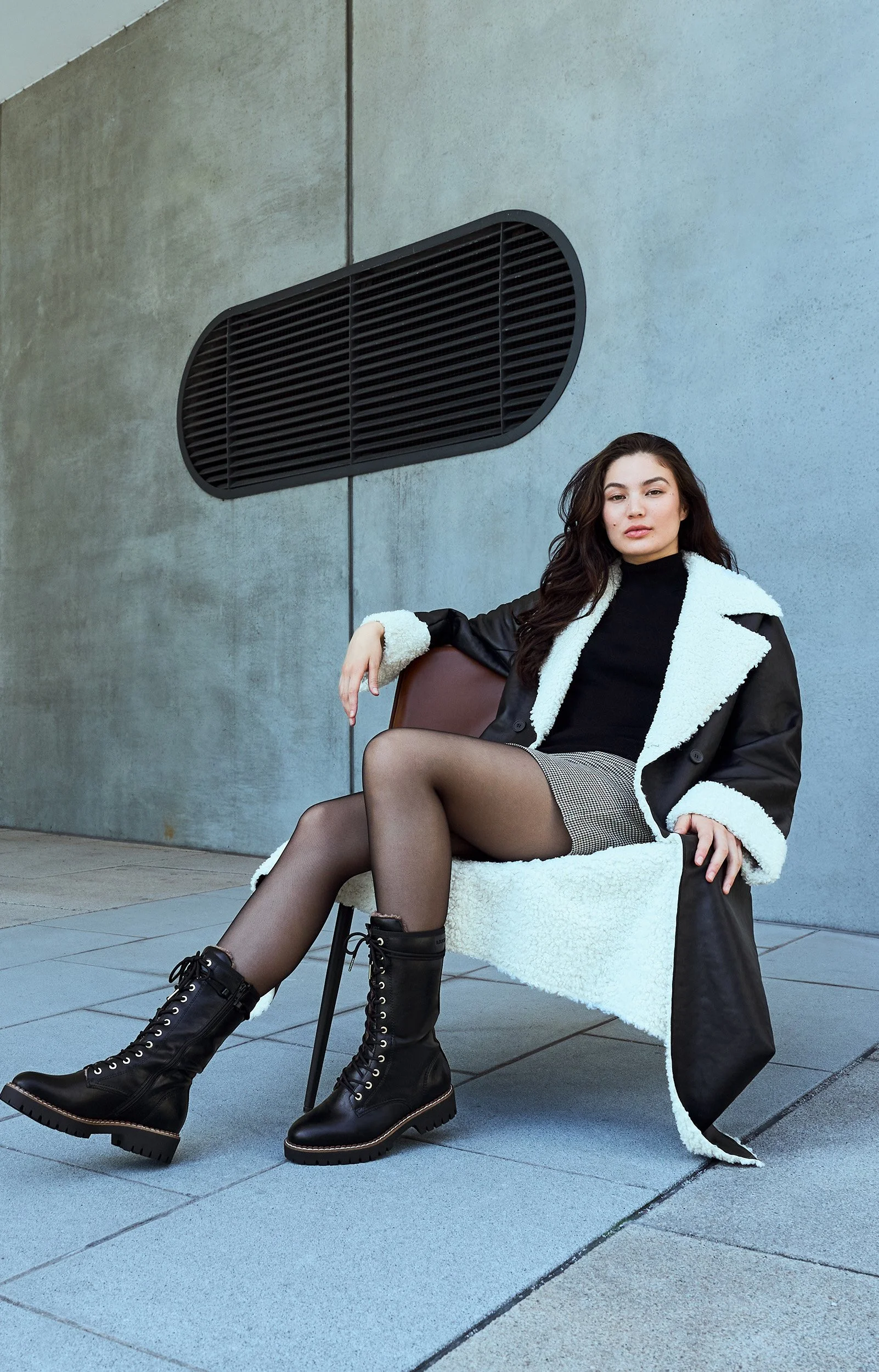 Junge Frau sitzt auf einem modernen Stuhl, trägt einen schwarzen Pullover, eine graue Tasche, schwarze Lederstiefel und einen schwarzen Mantel mit weißem Teddyfutter, vor einer grauen Betonwand.