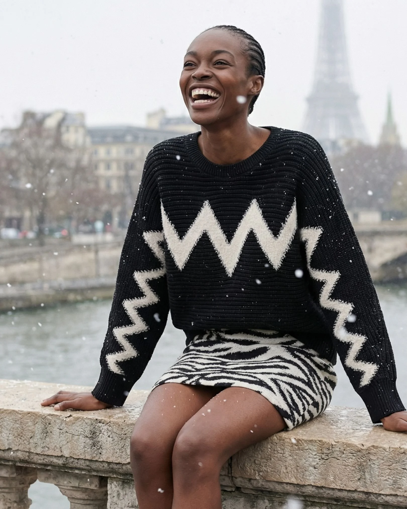 Eine lachende Frau sitzt auf einer Steinmauer an einem Fluss in Paris, während Schneeflocken fallen, mit der Eiffelturm im Hintergrund.