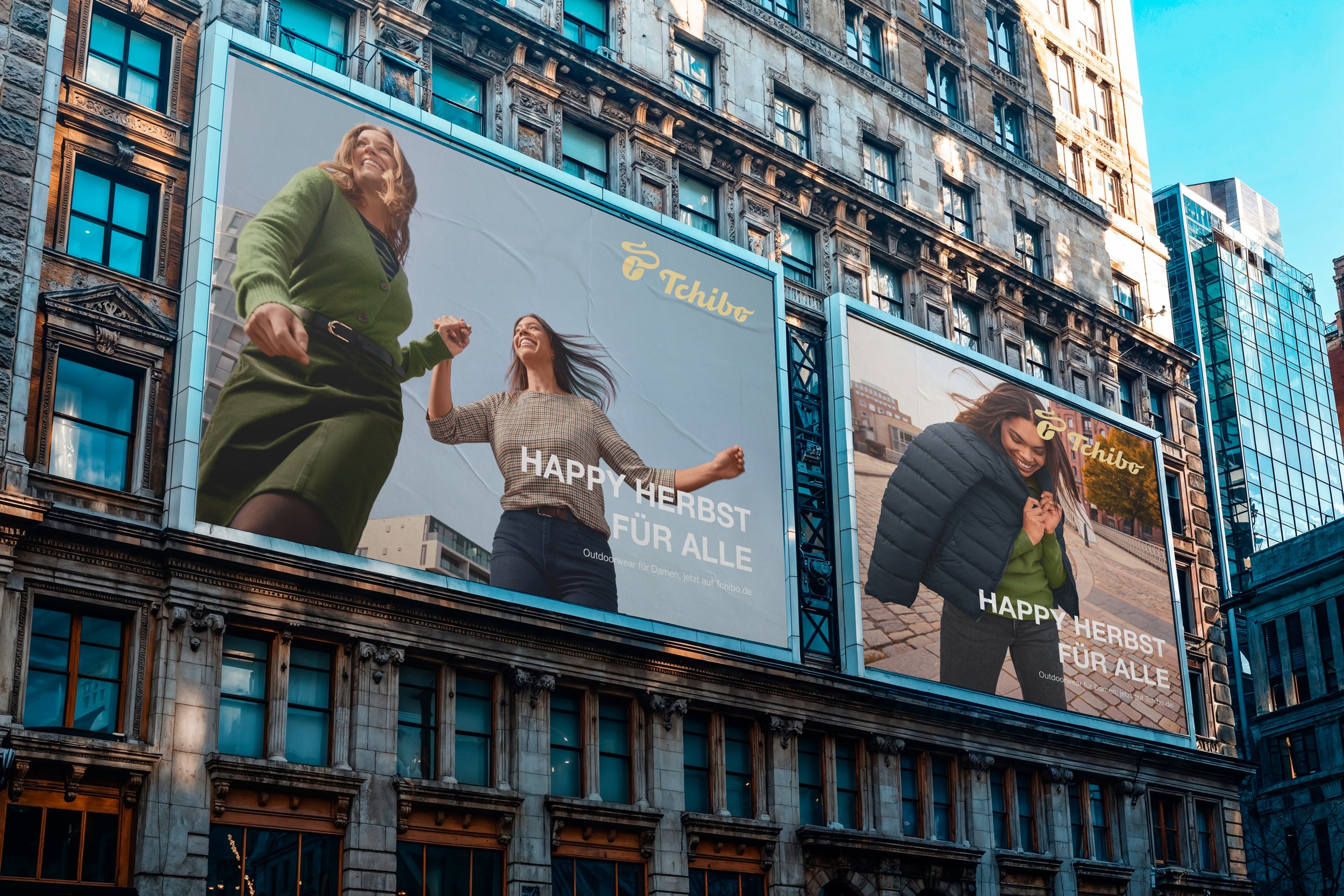 Zwei Werbeflächen auf einem Stadtgebäude mit Frauen, die lachen und sich freuen, Werbung für Tchibo, mit der Botschaft "Frohes Herbst für alle" auf Deutsch.