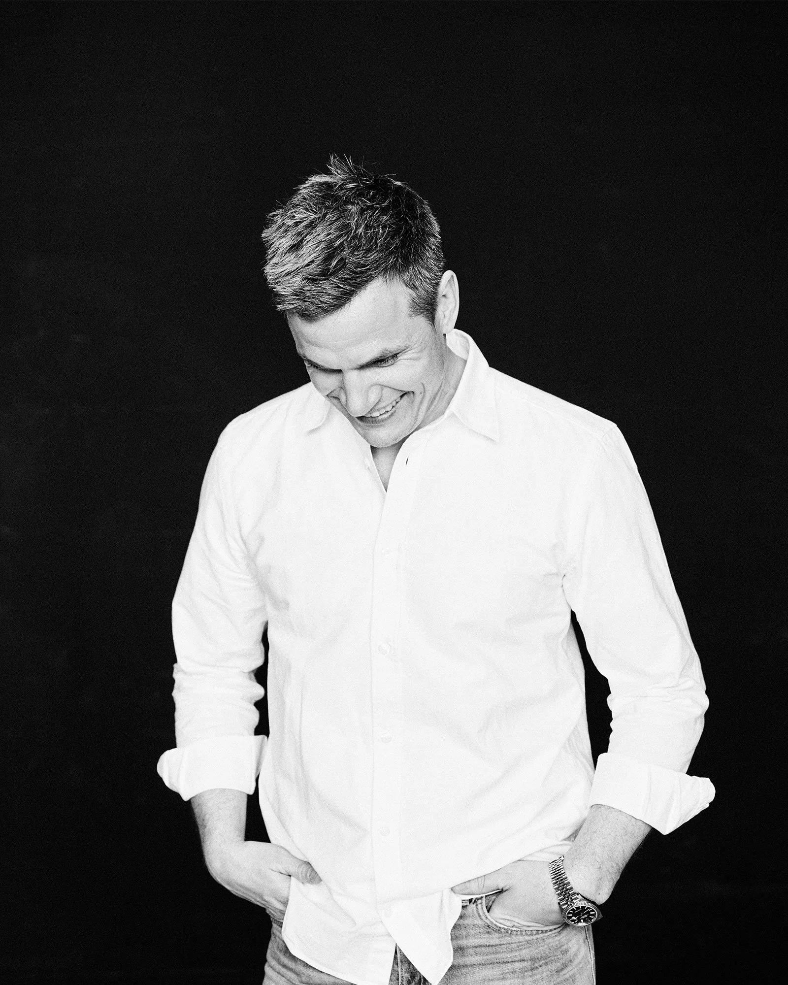 Schwarz-weiß Foto eines lachenden jungen Mannes in weißem Hemd, mit Händen in den Taschen, vor dunklem Hintergrund.