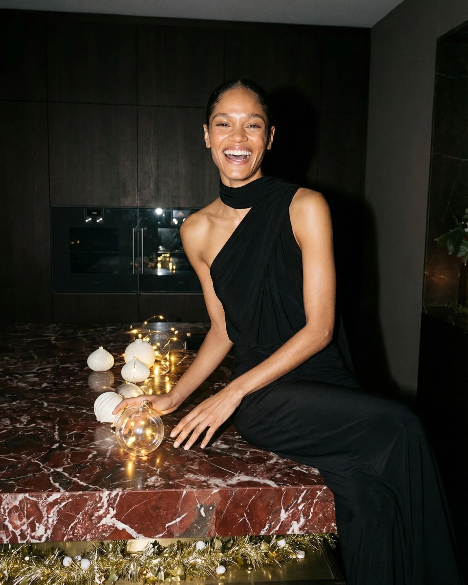 Eine lachende Frau in einem schwarzen Abendkleid sitzt an einem Marmortisch mit Weihnachtsdekoration und glitzernden Kugeln.