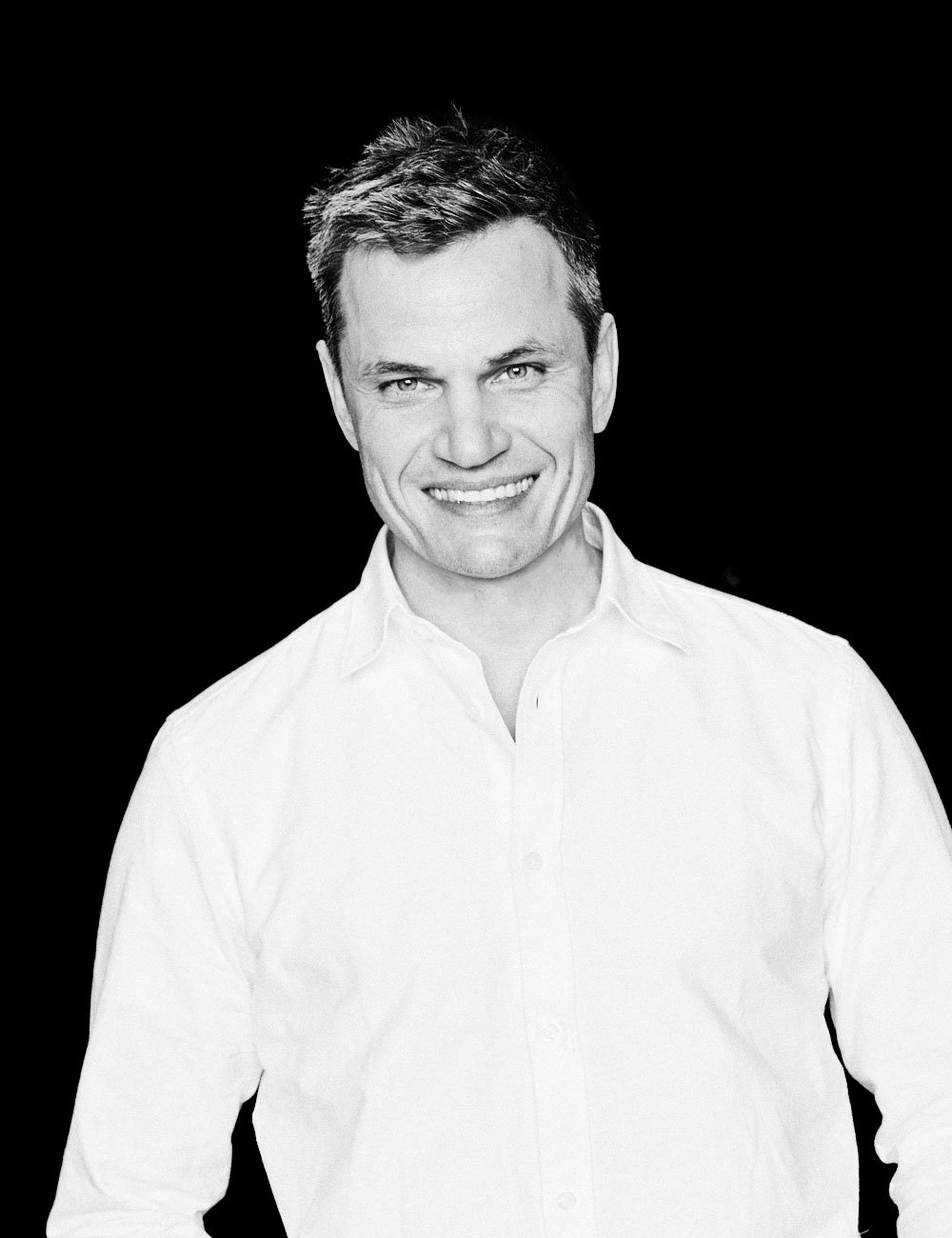 Schwarz-weiß Portrait eines lächelnden Mannes in weißem Hemd vor schwarzem Hintergrund.